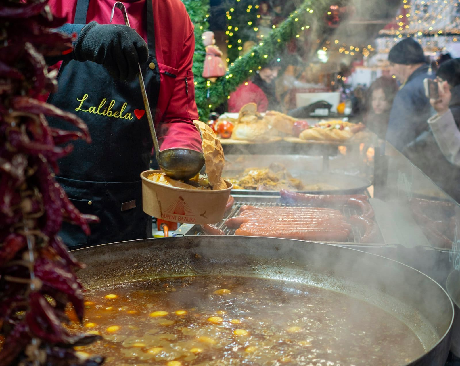 Straßenimbissverkäufer serviert traditionellen ungarischen Eintopf aus einem großen Topf auf einem Outdoor-Markt in Budapest mit Menschen und festlicher Dekoration im Hintergrund.