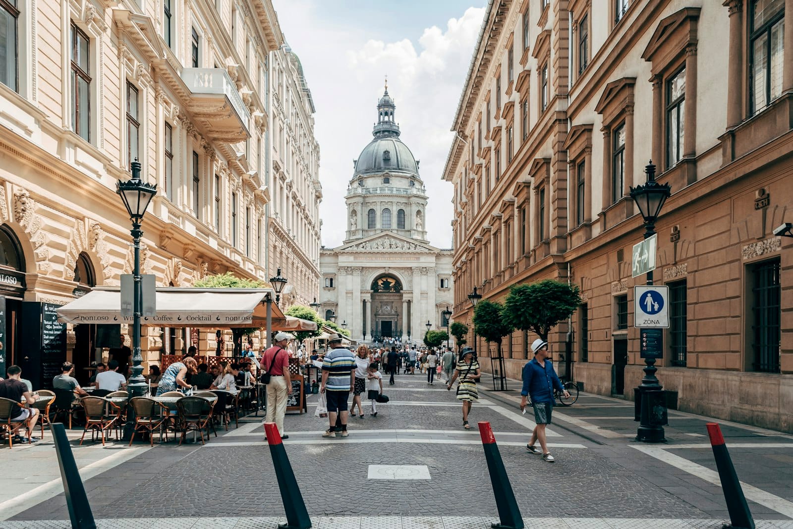 Straßenszene im Zentrum von Budapest mit Menschen beim Essen im Freien in Cafés, historischer Architektur und der Basilika Sankt Stephan im Hintergrund.