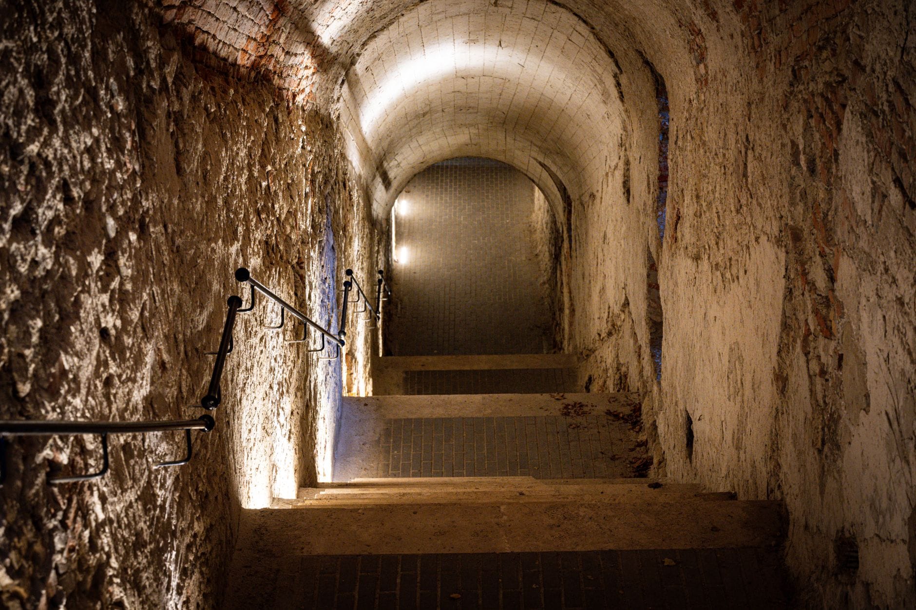 Steinstufen führen hinab in einen beleuchteten, gewölbten unterirdischen Gang mit rauen, historischen Wänden, der an verborgene Tunnel oder Bunker unter einer Stadt erinnert.