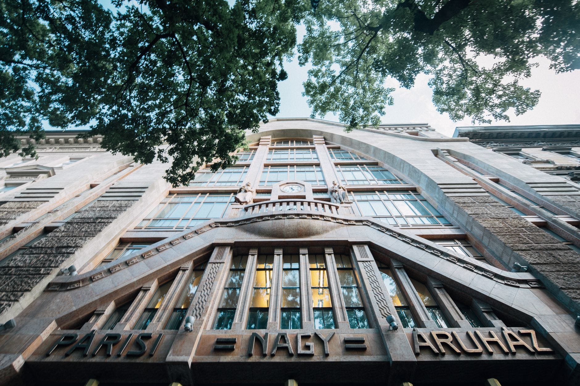 Blick nach oben auf die kunstvolle Jugendstil-Fassade des Párisi Nagy Áruház in Budapest, gerahmt von Ästen.