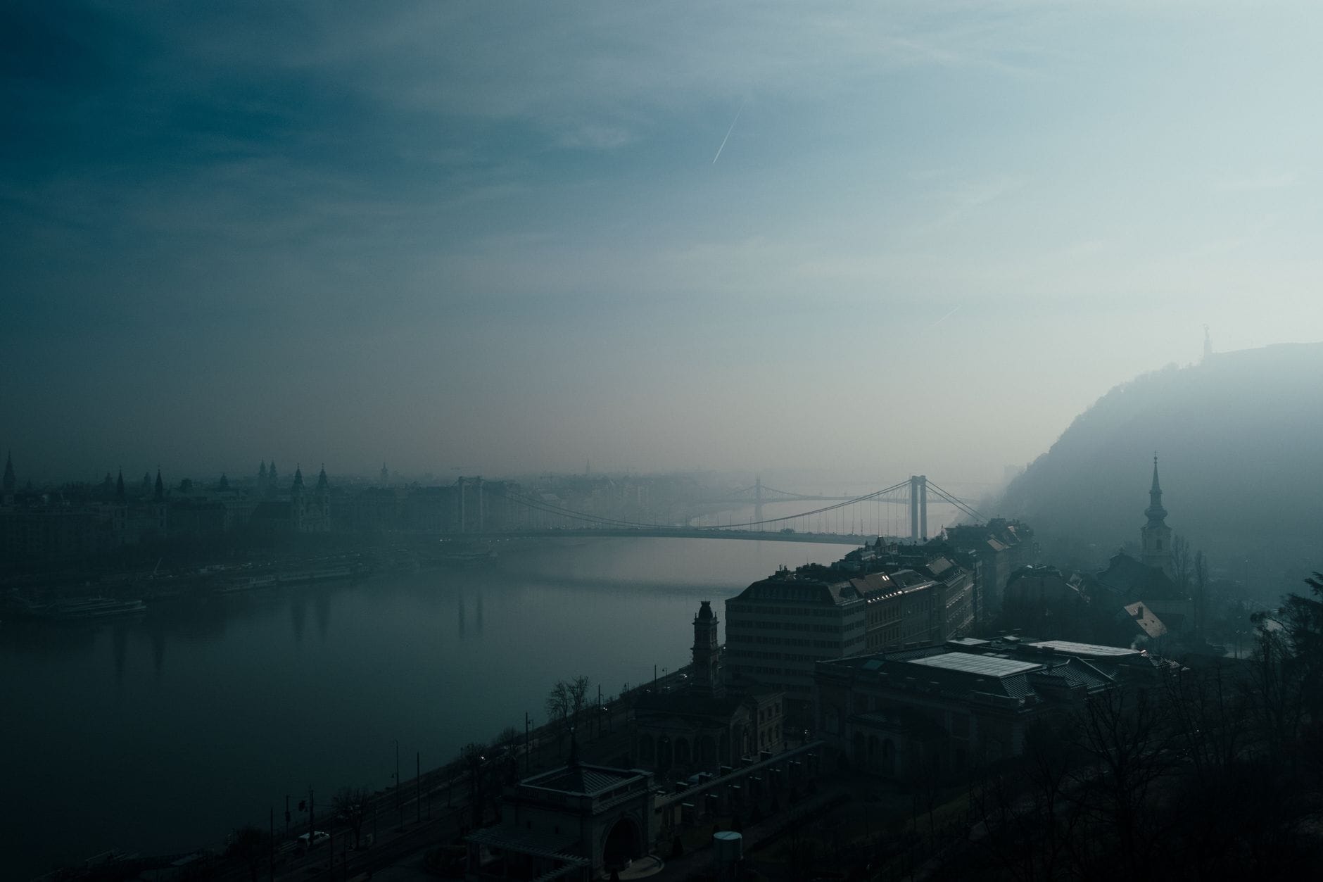 Stimmungsvolle, neblige Aussicht auf die Skyline von Budapest und den Fluss, mit einer Brücke über die Donau und historischen Gebäuden entlang des Ufers.