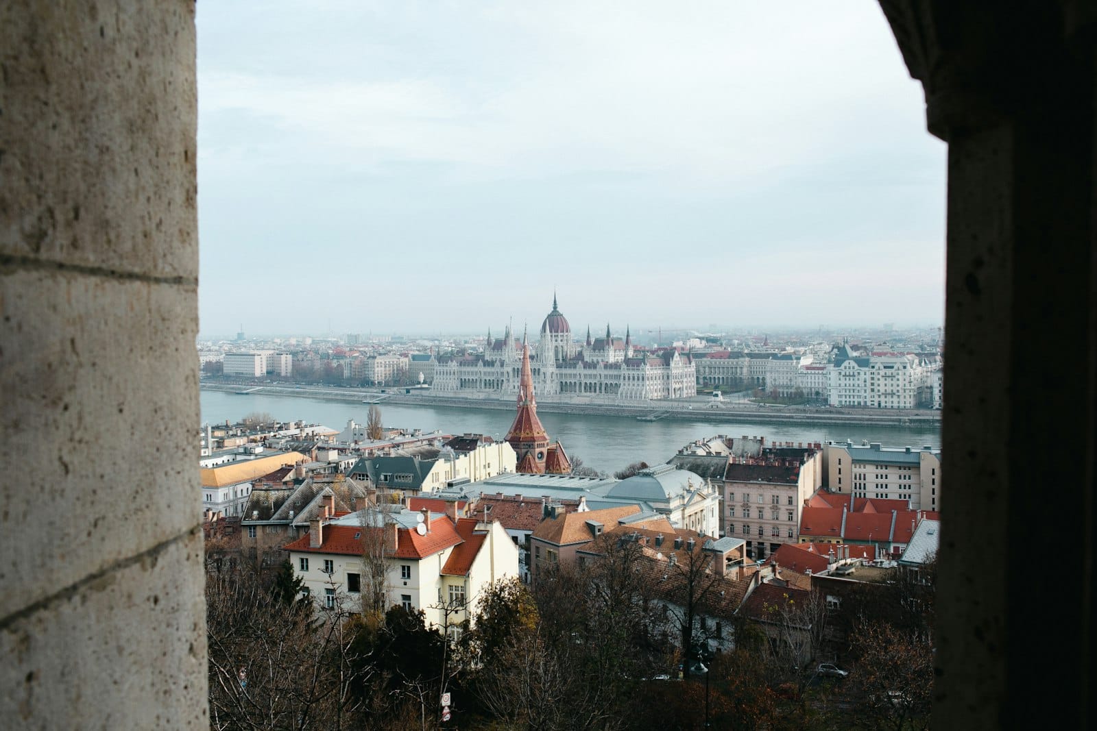 Blick auf Budapest mit bedecktem Himmel, kahlen Winterbäumen und dem Parlamentsgebäude auf der anderen Seite der Donau, durch Steinbögen gesehen.
