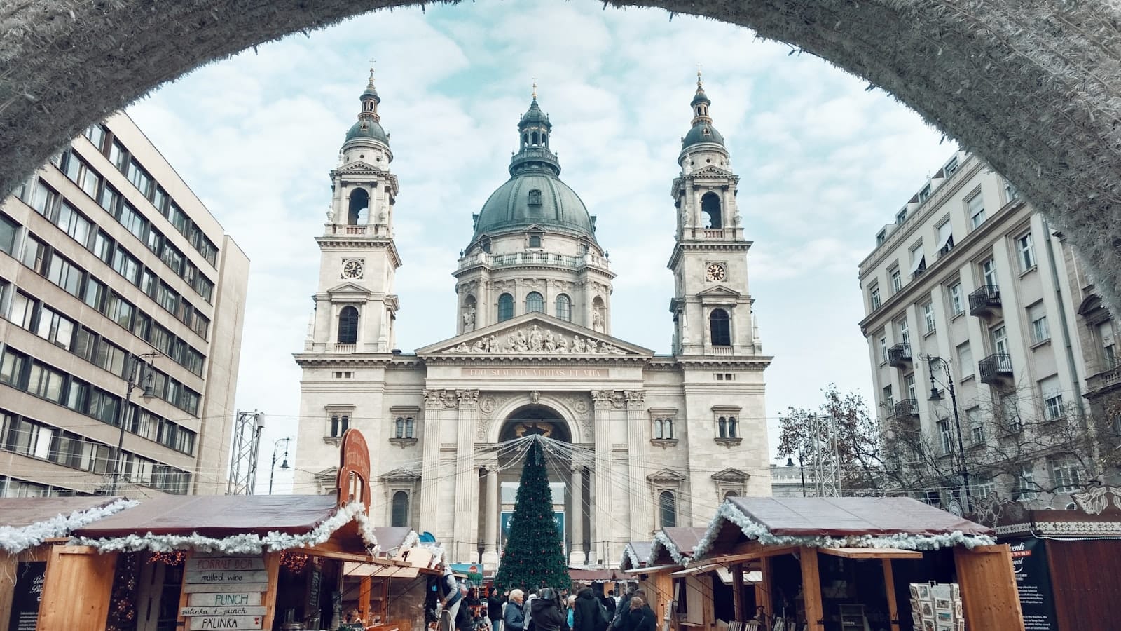 Die Basilika St. Stephan mit Weihnachtsmarktständen und einem großen Weihnachtsbaum, gesehen durch einen festlichen Torbogen in Budapest an einem Wintertag.