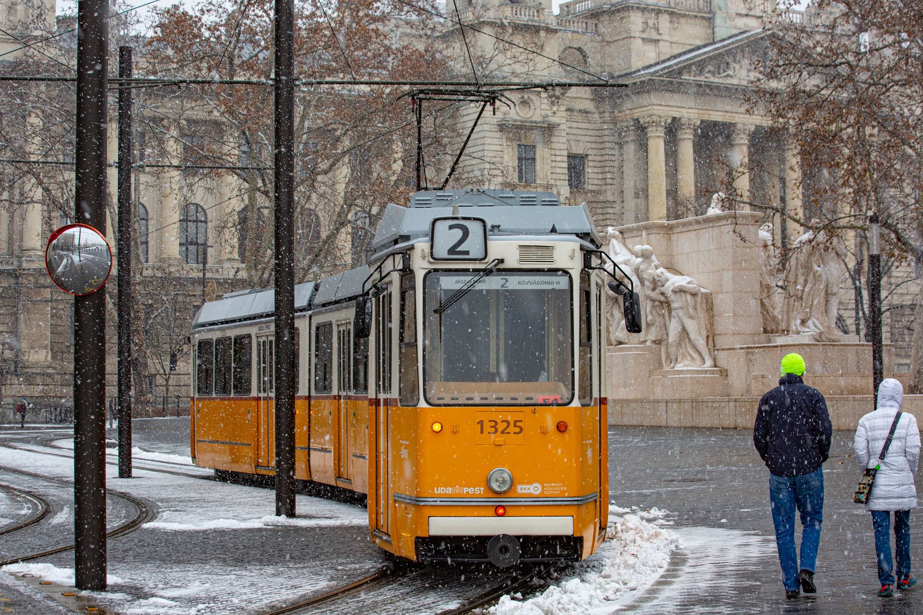 Gelbe Straßenbahn in Budapest im Winterschnee, mit zwei vorbeilaufenden Personen und historischer Architektur im Hintergrund.