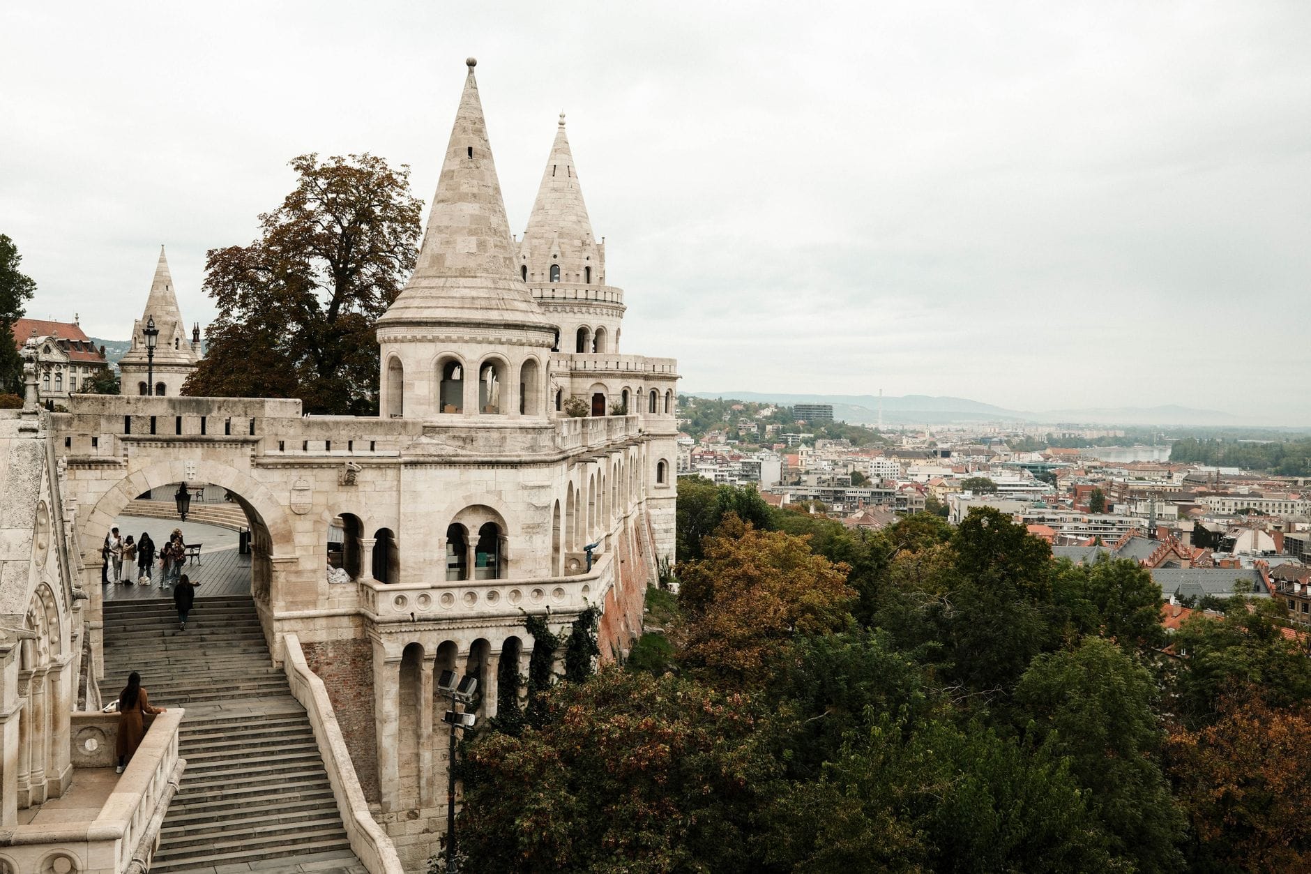 Bulutlu bir günde Budapest'teki Balıkçı Kalesi'nin kulecikleri, taş kemerleri ve arka planda şehir manzarasıyla geniş görünümü.