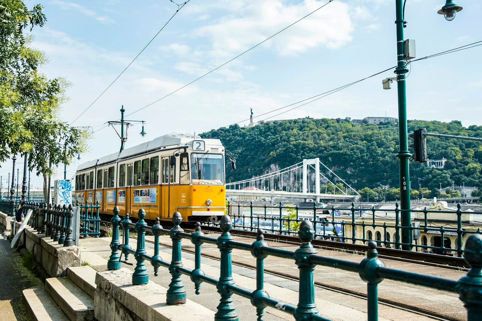 Güneşli bir günde arka planda tepeler ve köprü ile Tuna Nehri boyunca ilerleyen sarı Budapest tramvayı