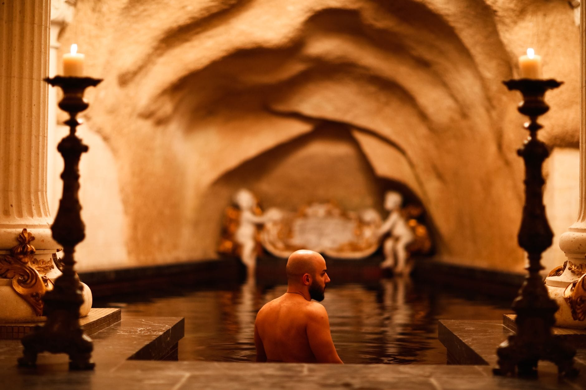 Ein Mann entspannt sich in einem prunkvollen Thermalbad mit gewölbter Steindecke, brennenden Kerzen und barock inspiriertem Dekor, das die Luxus-Spa-Kultur von Budapest heraufbeschwört.