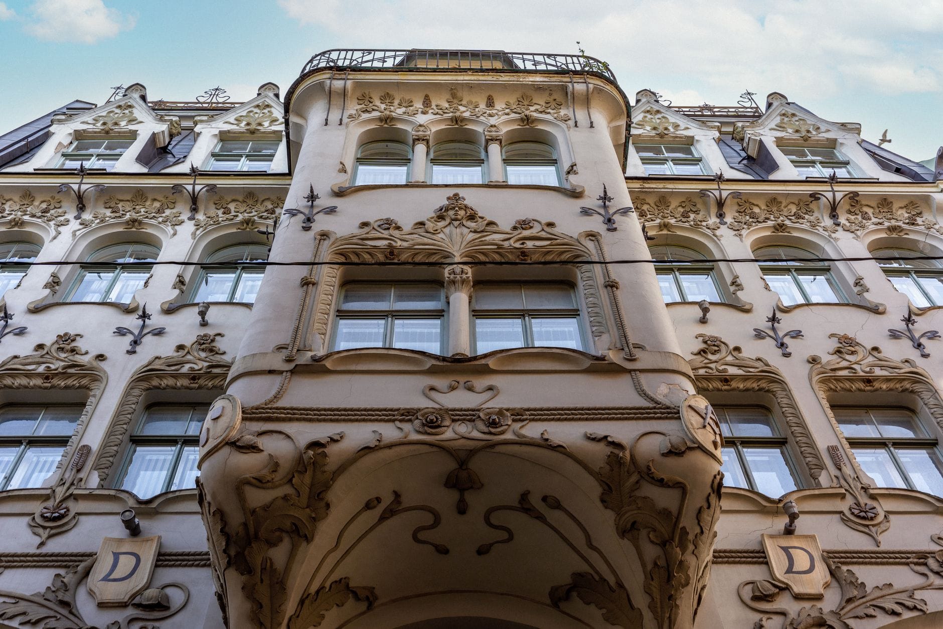 Verzierte Fassade eines Jugendstilgebäudes in Budapest mit aufwendigem Stuck und romantischen Architekturdetails, von unten fotografiert mit blauem Himmel.