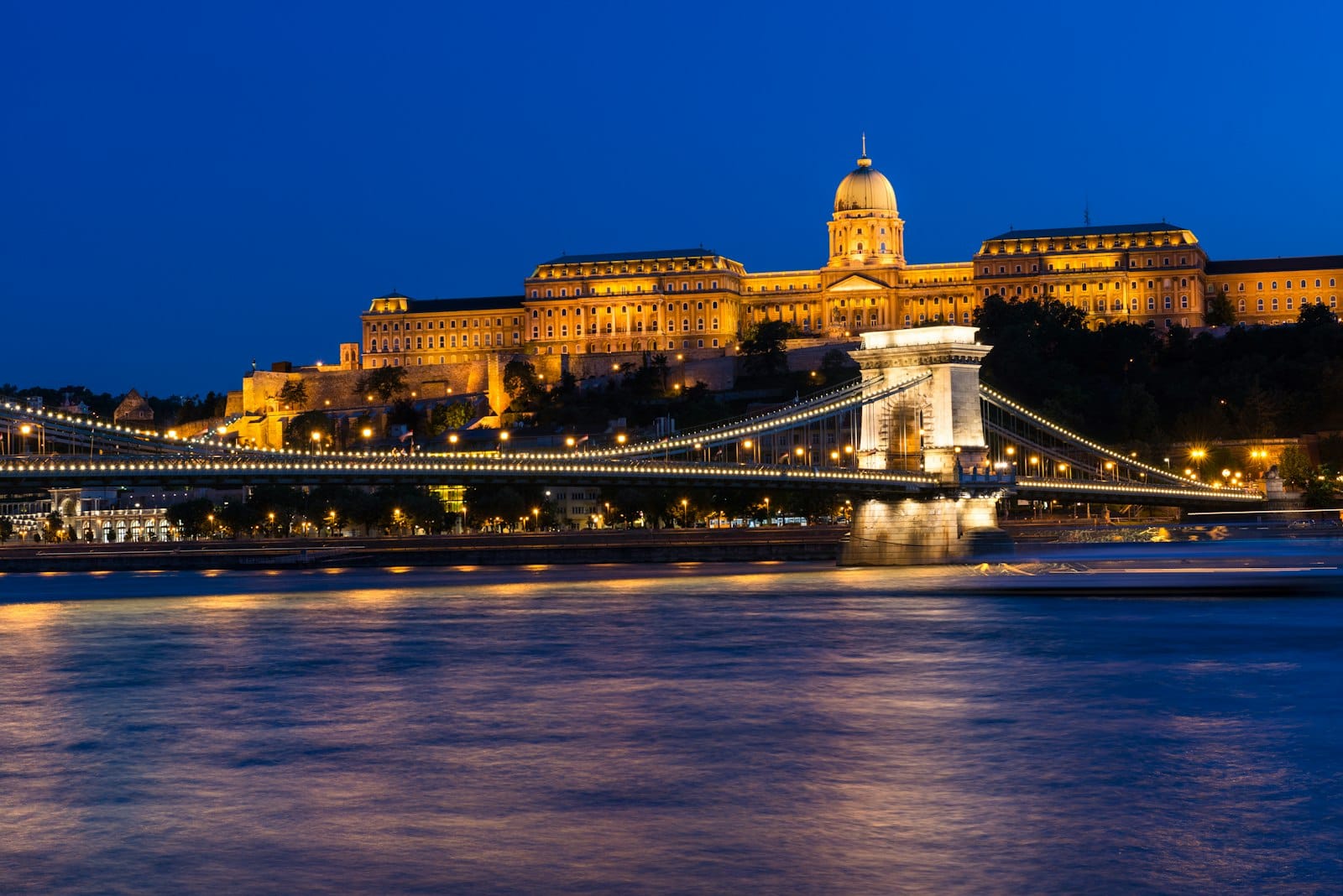 Night view of Budapest’s Chain Bridge illuminated over the Danube River with Buda Castle glowing in the background under a deep blue sky.