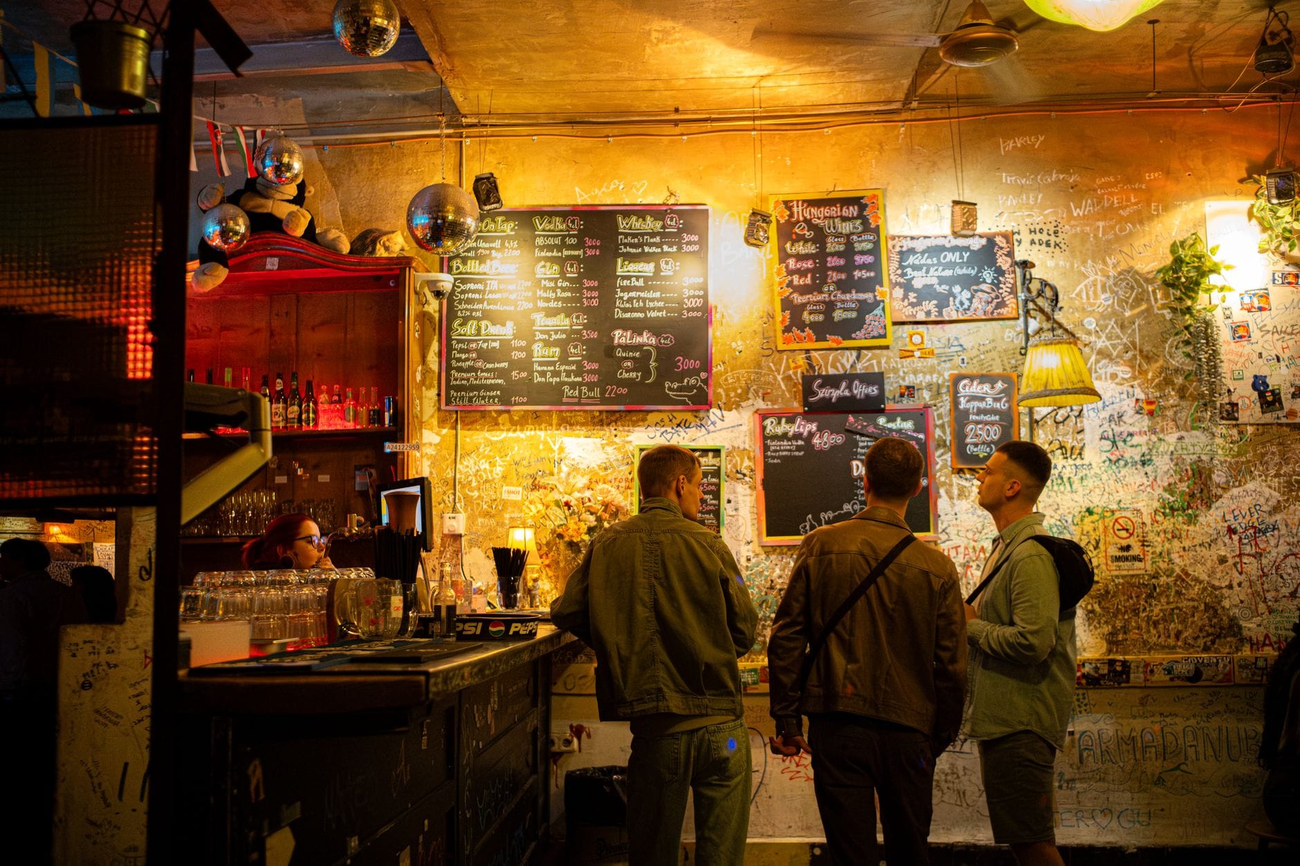 Interno di un bar eccentrico con pareti coperte di graffiti, arredamento eclettico e persone in piedi vicino a un bancone con i menu delle bevande a Budapest.