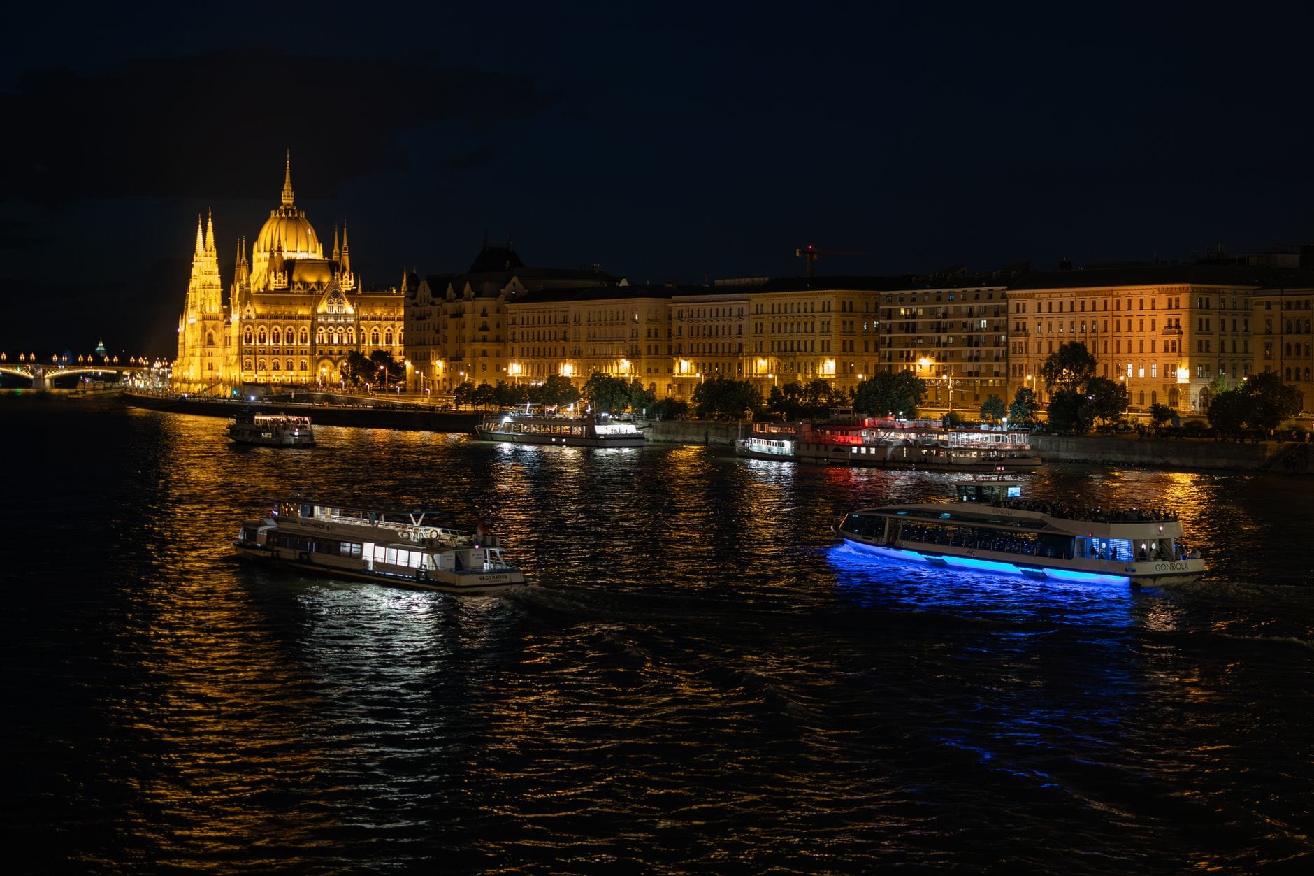 Vista notturna del Parlamento di Budapest e del lungofiume con più barche illuminate che navigano sul Danubio.