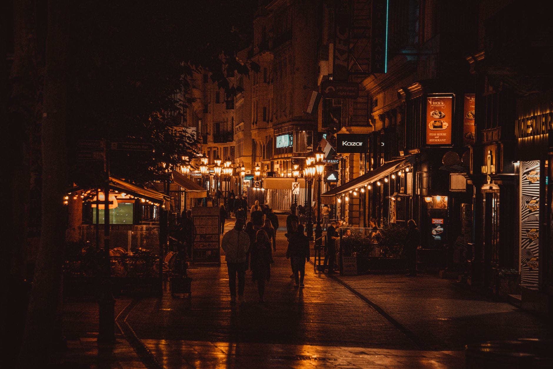 Scena notturna di una vivace strada di Budapest costellata di bar e luci brillanti, con persone che passeggiano e vetrine illuminate che creano un'atmosfera di vibrante vita notturna.