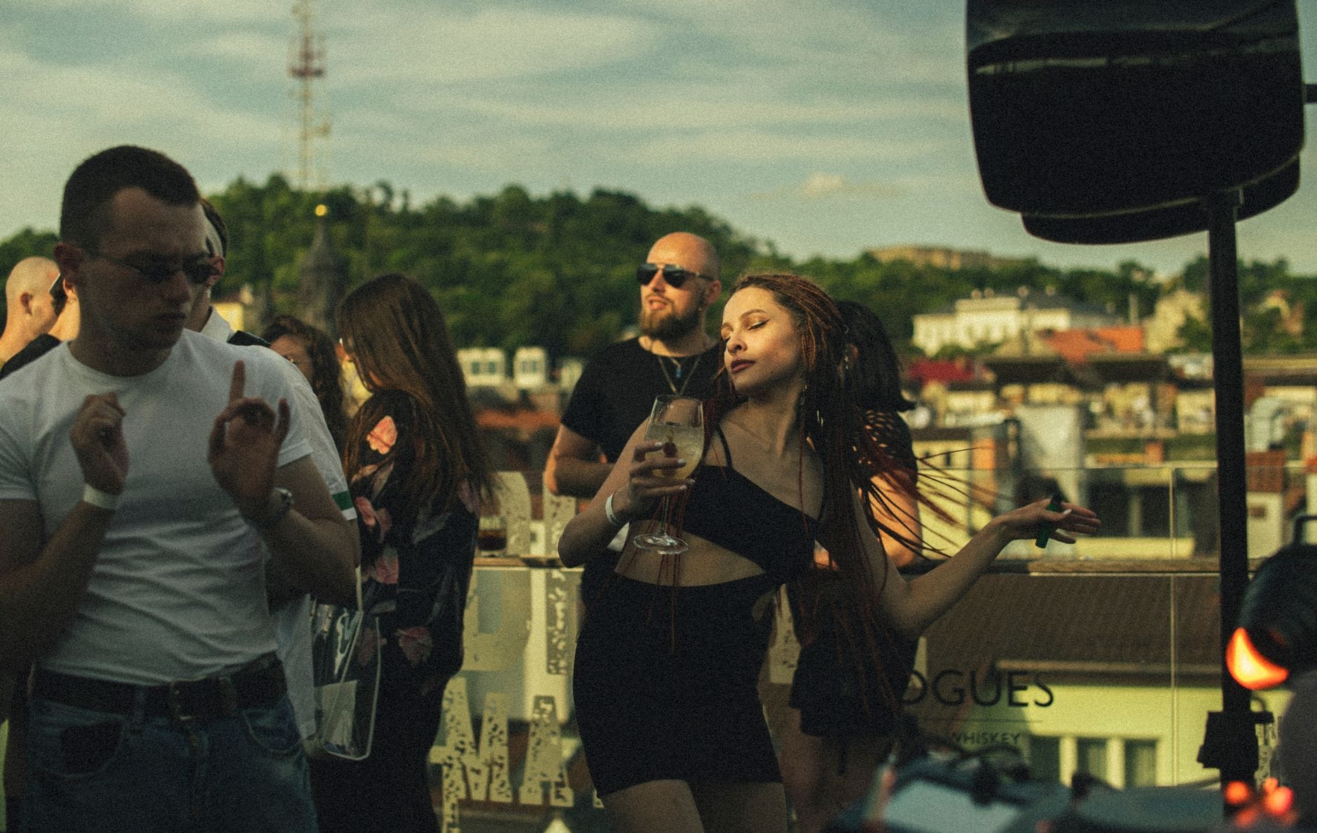Giovani adulti che si godono drink e ballano in un rooftop bar con il panorama di Budapest e le colline sullo sfondo in una giornata di sole.