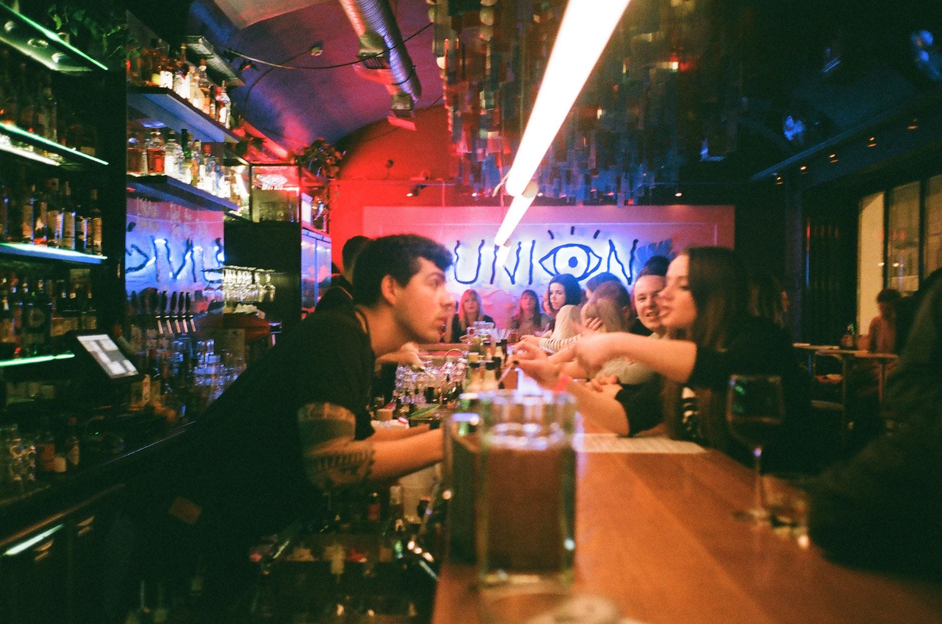 Animado bar abarrotado con un bartender sirviendo bebidas a jóvenes en un local ecléctico y colorido con luces de neón y ambiente vibrante.