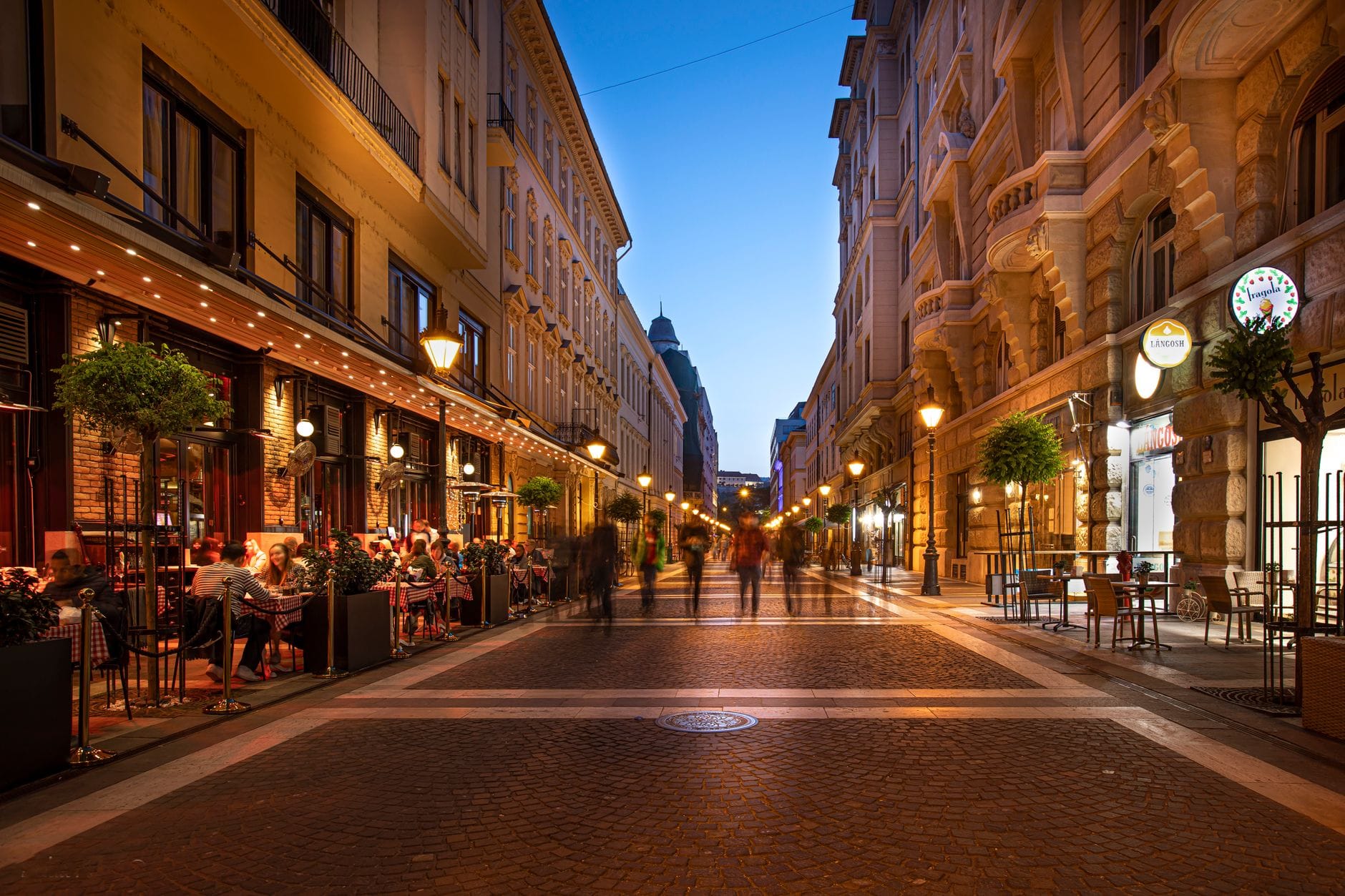 Escena nocturna en una animada calle de Budapest con personas cenando al aire libre y otras paseando, iluminada por cálidas luces, mostrando la vibrante atmósfera estacional y el flujo de la multitud.