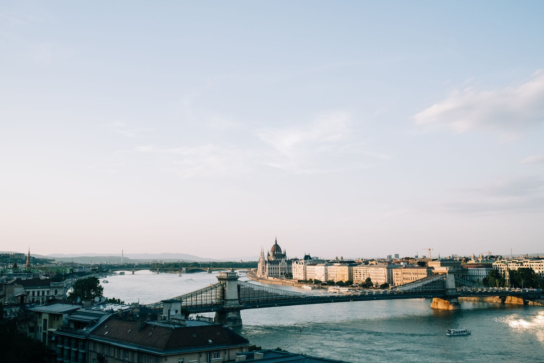 Tagesansicht des Budapester Parlaments und der Kettenbrücke über die Donau mit klarem Himmel und Stadtgebäuden entlang der Uferpromenade.