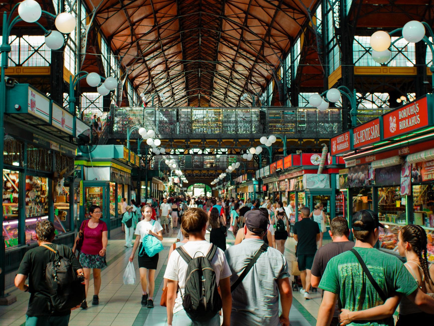 Innenansicht der Großen Markthalle in Budapest mit Besuchern, die zwischen bunten Ständen mit lokalen Lebensmitteln und Souvenirs schlendern – unter einem prachtvollen Dach aus Eisen und Glas.