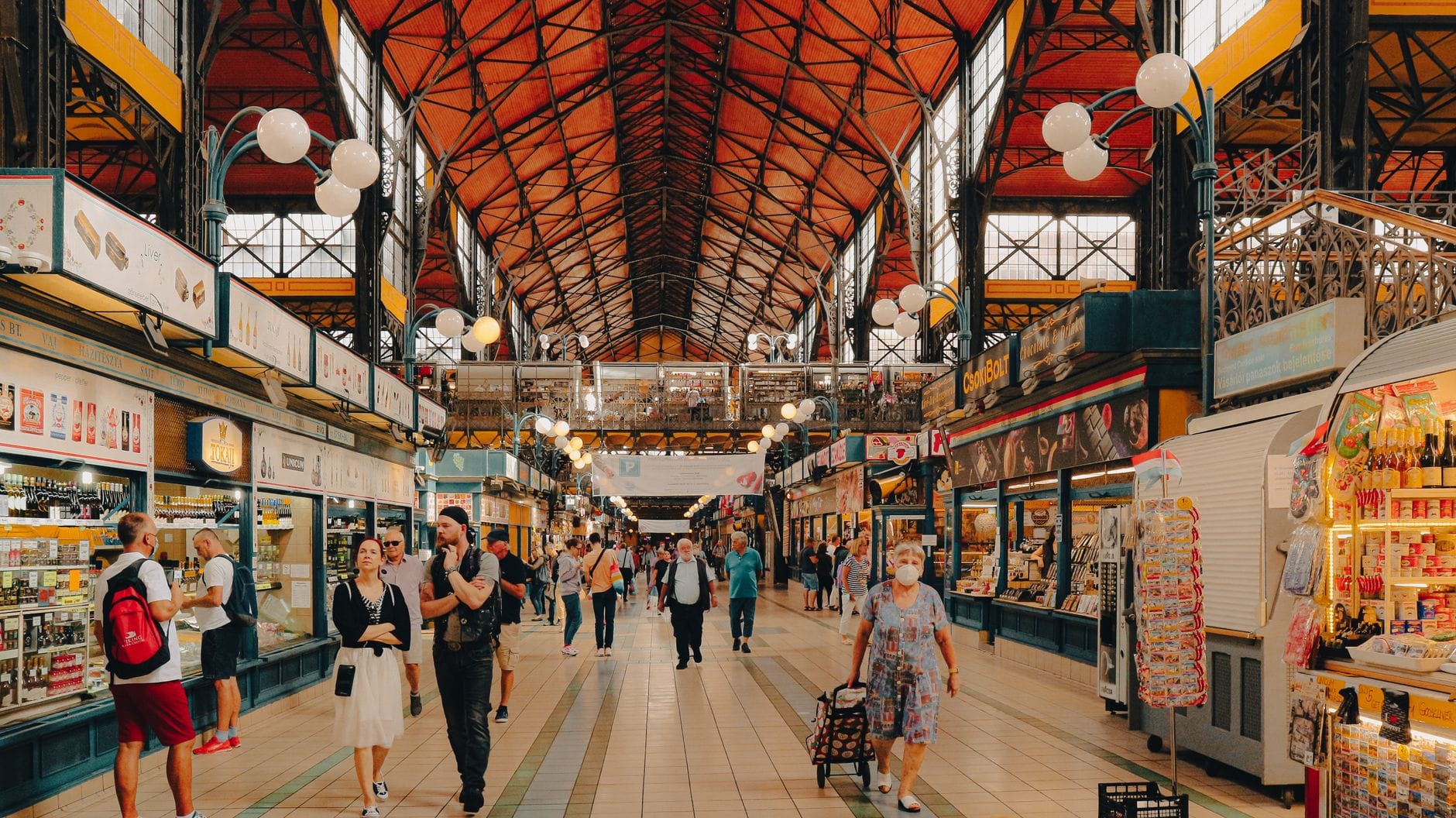 Weitwinkelansicht des Inneren der Großen Markthalle Budapest mit einkaufenden Menschen, Ständen auf beiden Seiten und einem hohen Gewölbedach mit schmiedeeisernen Details.
