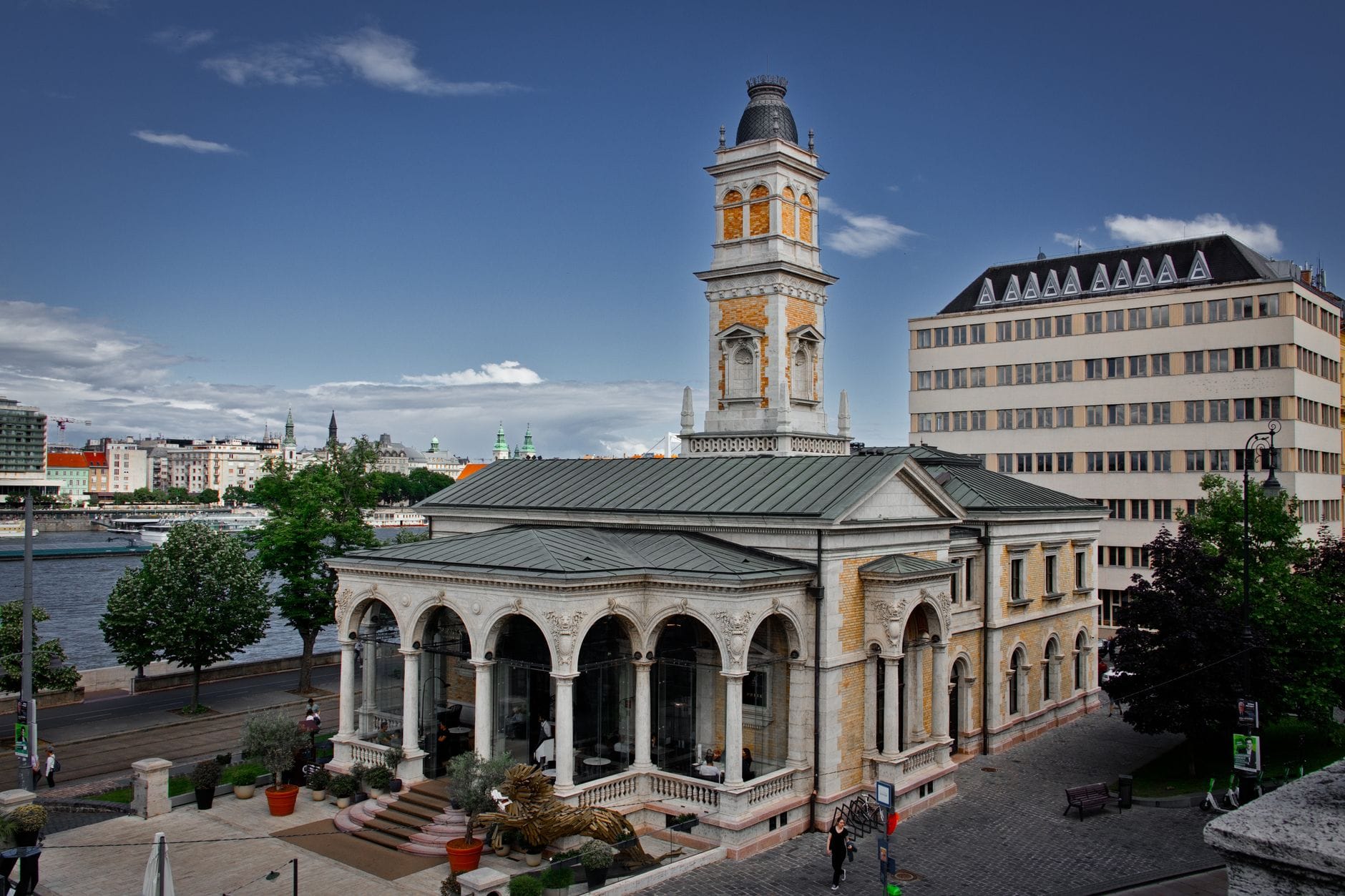 Historisches Gebäude mit Bogenfenstern und einem Uhrenturm, am Flussufer in Budapest mit Fußgängern in der Nähe.