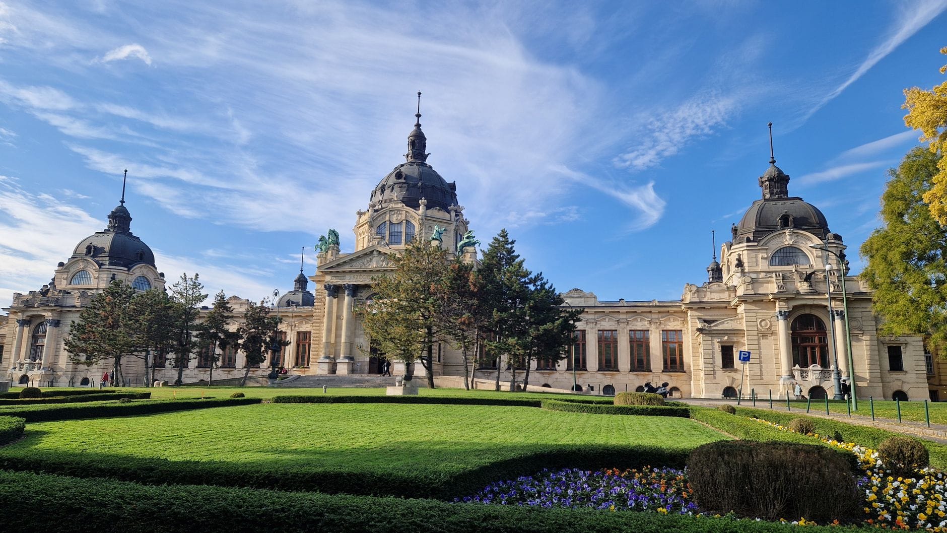 Széchenyi Thermalbad in Budapest an einem klaren Tag mit sehr wenigen Menschen davor, mit beeindruckender Fassade und weitläufigem Garten.