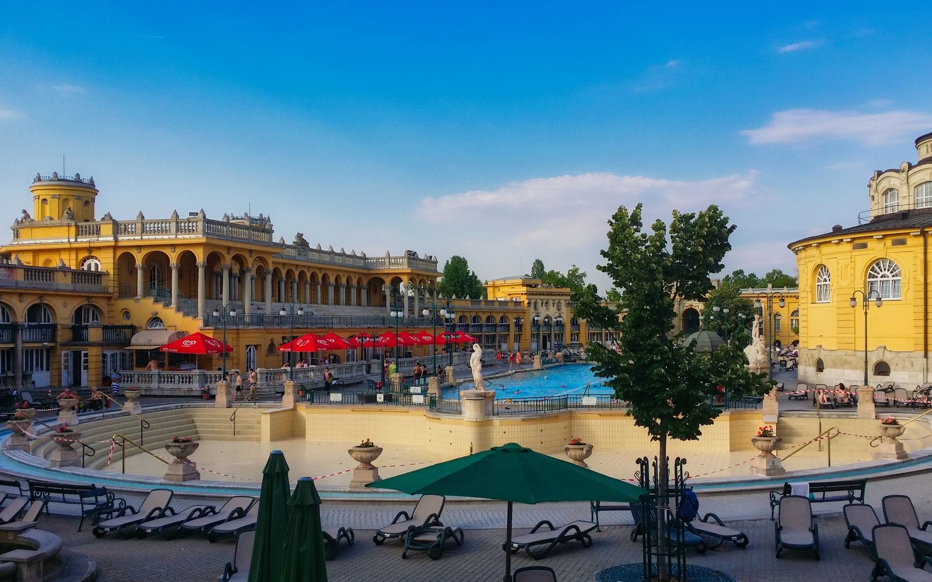 Weitwinkelansicht des Innenhofs der Széchenyi-Therme mit gelben neobarocken Gebäuden, Außenpools, roten Sonnenschirmen und entspannenden Menschen.