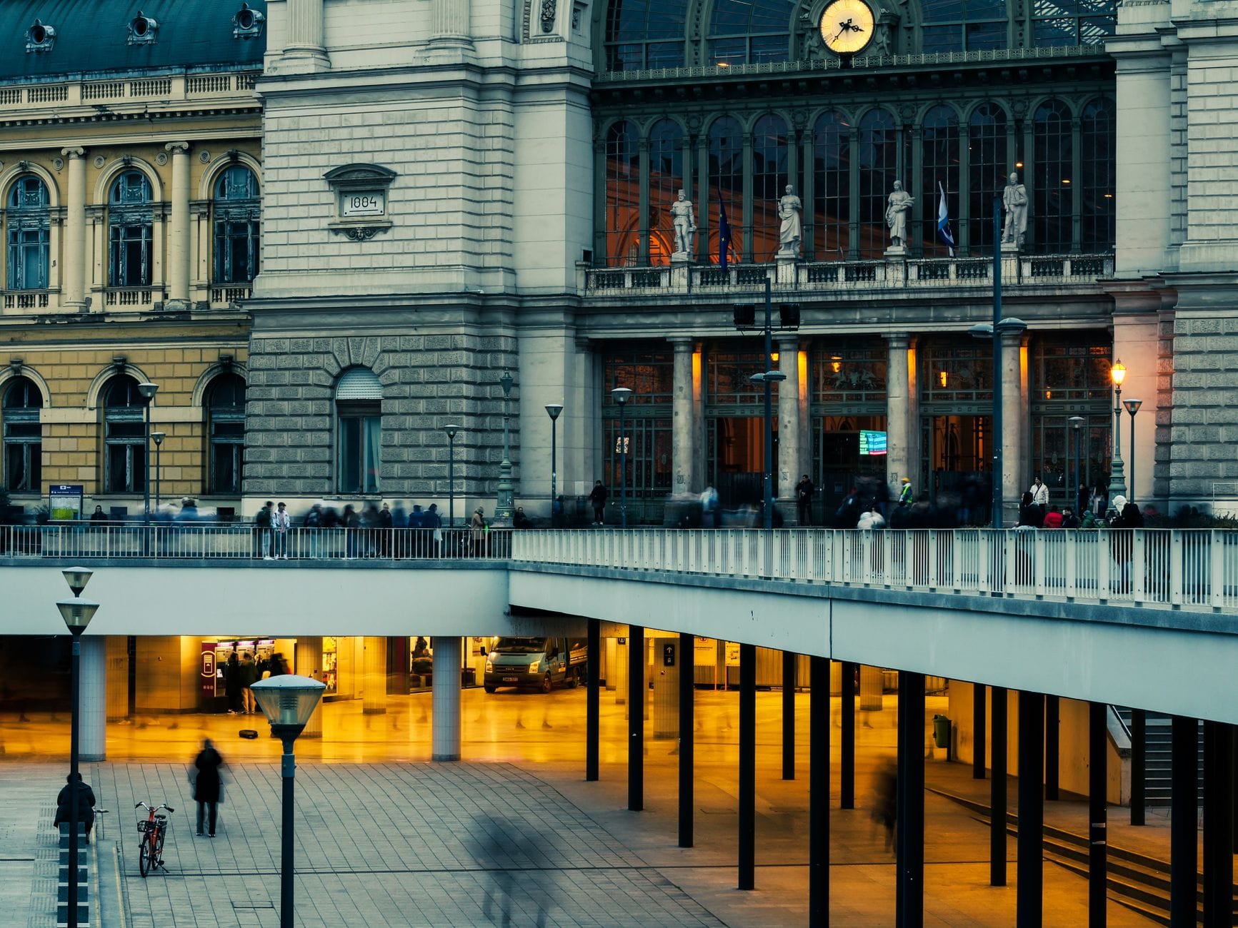 Widok zewnętrzny dworca kolejowego Budapest Keleti z jego okazałą fasadą ze szkła i kamienia, zegarem i ludźmi spacerującymi na pierwszym planie.