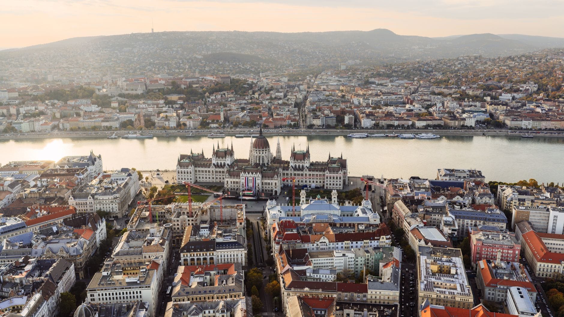 Una spettacolare vista aerea di Budapest con il Parlamento ungherese al centro, il Danubio, i tetti della città e le colline verdi sullo sfondo sotto un cielo dorato e soffuso.
