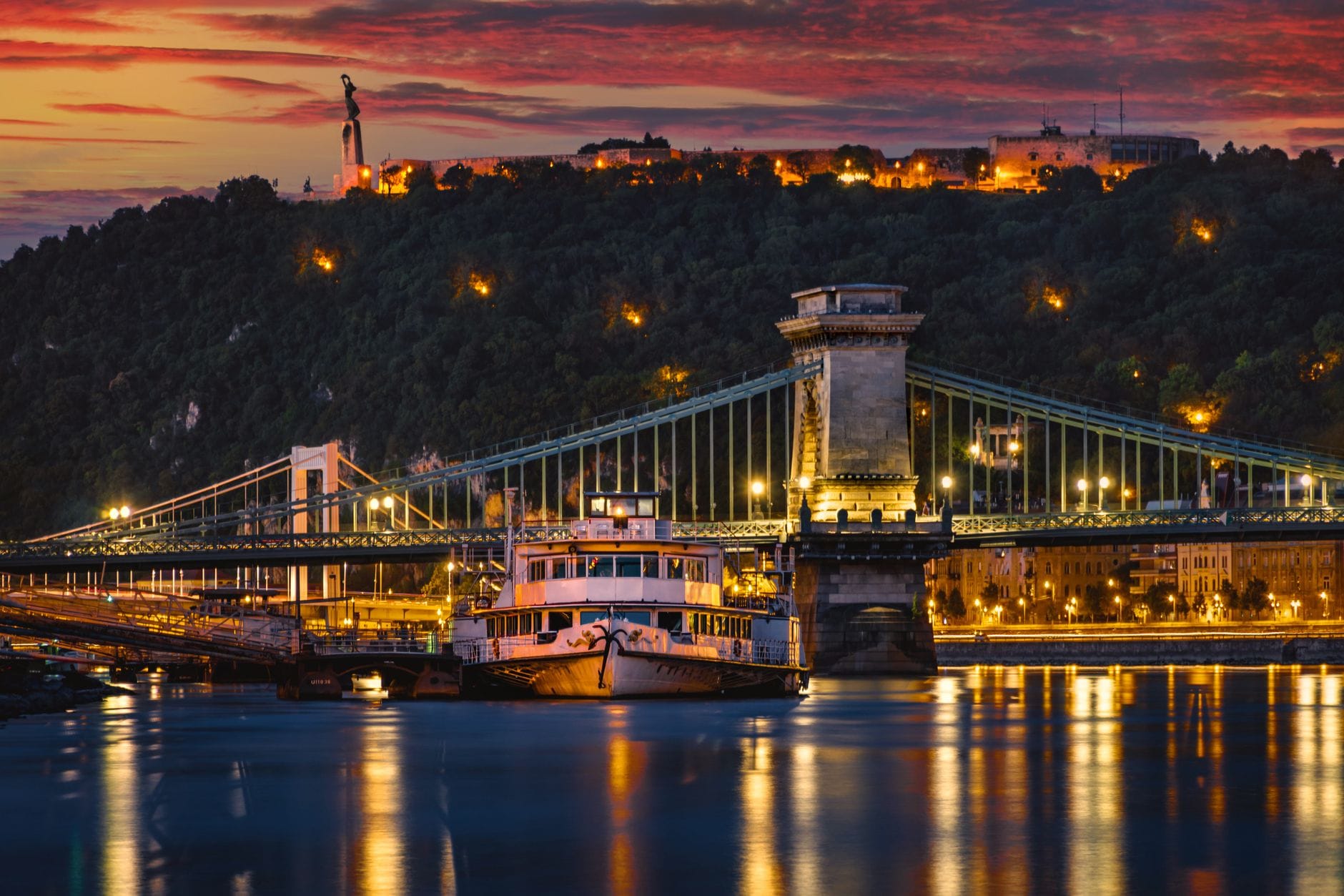 Vista notturna del Ponte delle Catene di Budapest sul Danubio con barche sull'acqua e le luci della città che brillano su uno spettacolare cielo al tramonto.