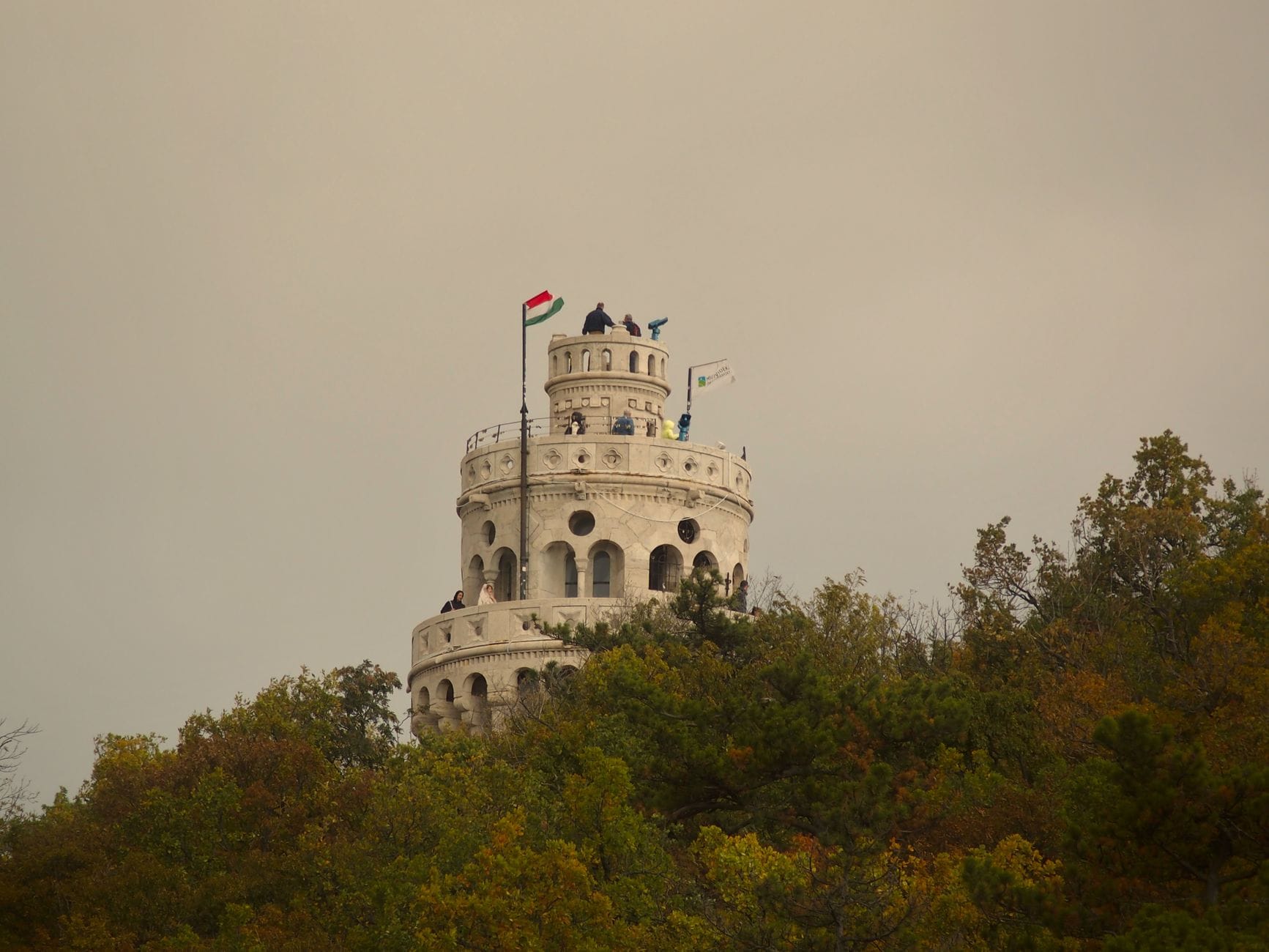 Torre di avvistamento in pietra bianca sulla cima di una collina boscosa, bandiera ungherese al vento, circondata da alberi autunnali sotto un cielo coperto