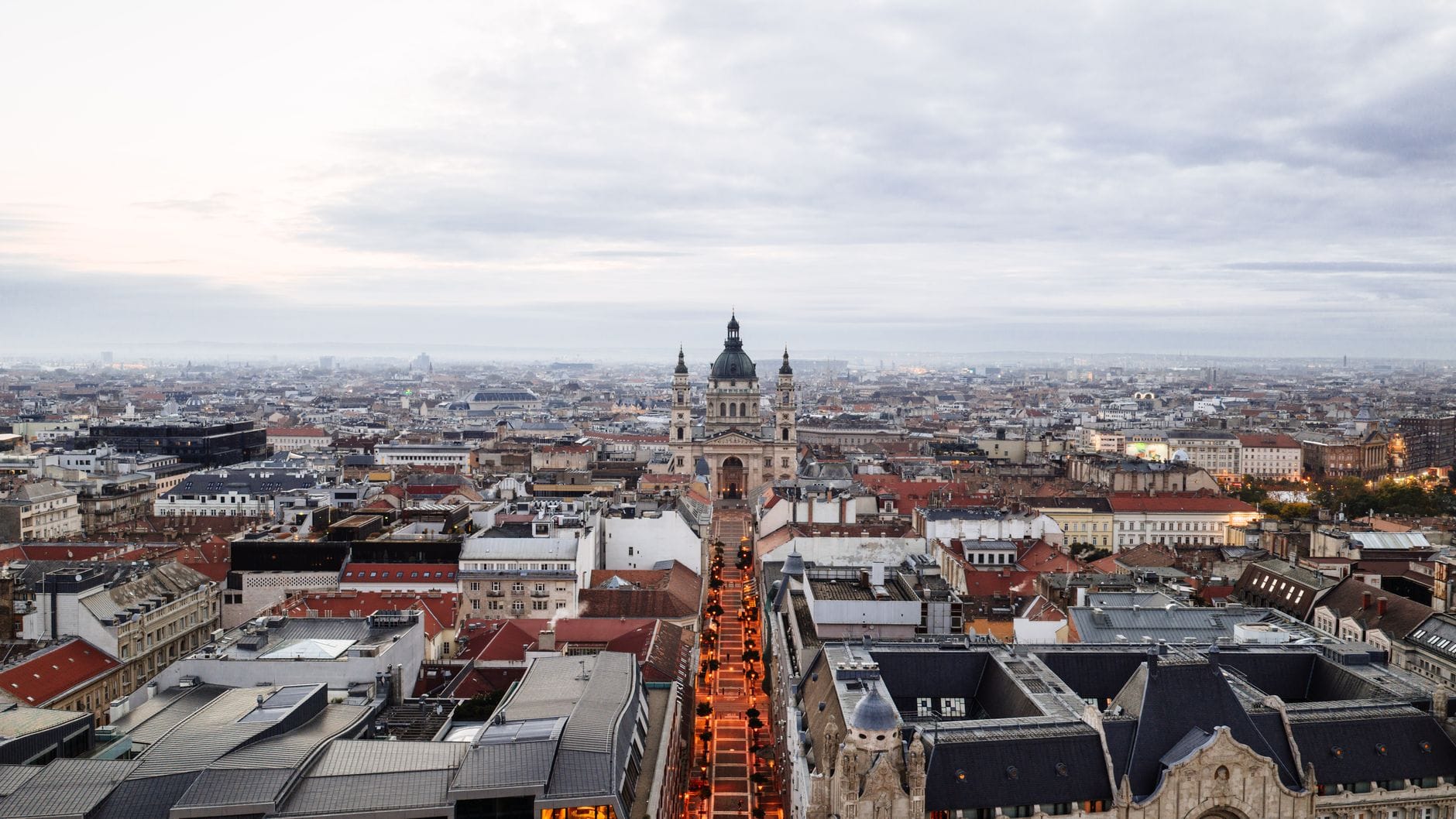 Ampia vista elevata del panorama urbano di Budapest con la Basilica di Santo Stefano al centro; vivaci profili dei tetti e ampie strade visibili sotto un cielo luminoso e nuvoloso.