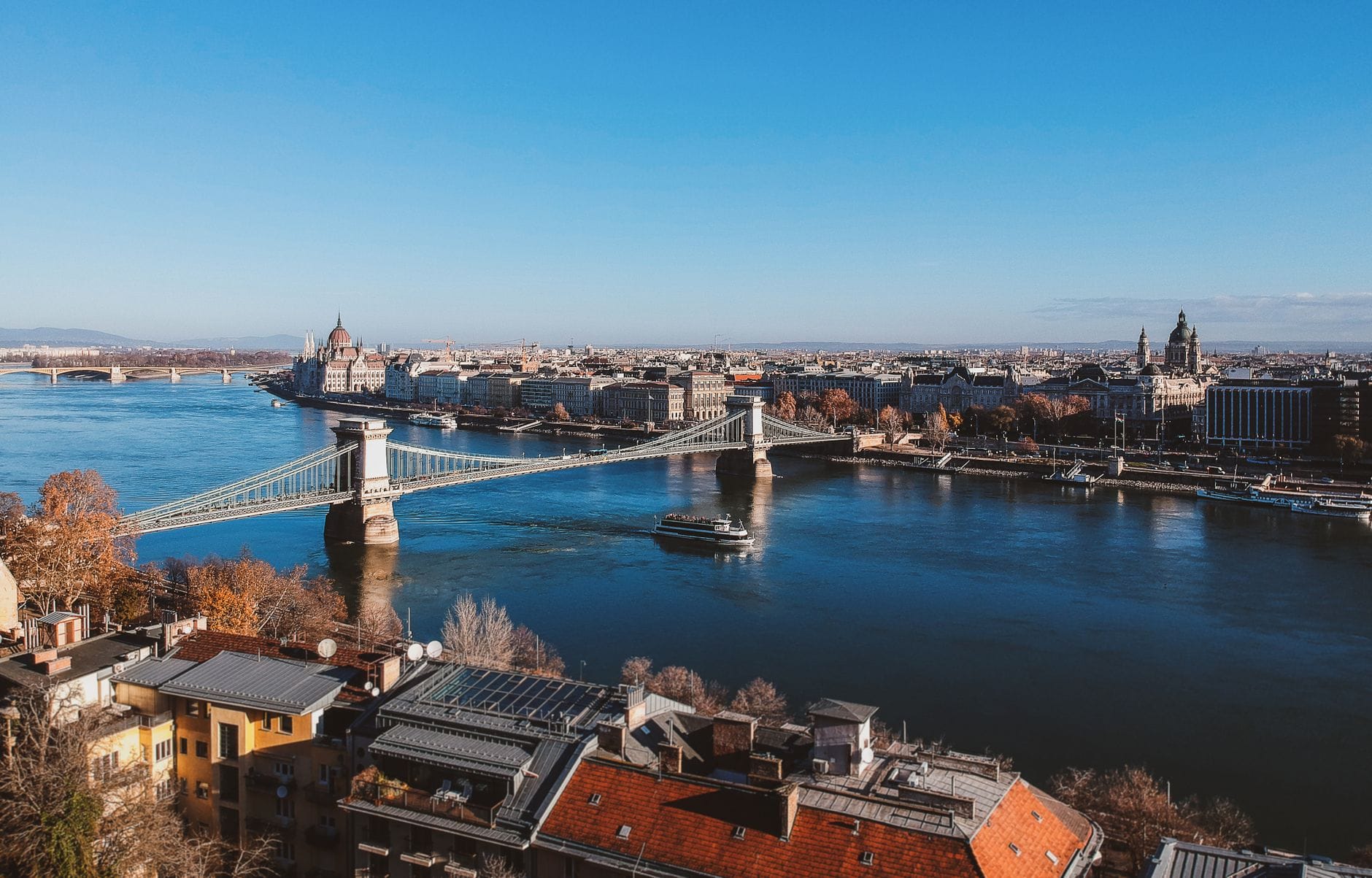 Pemandangan luas Budapest dengan Sungai Danube, Jembatan Rantai, kapal pesiar, dan landmark kota di bawah langit cerah.
