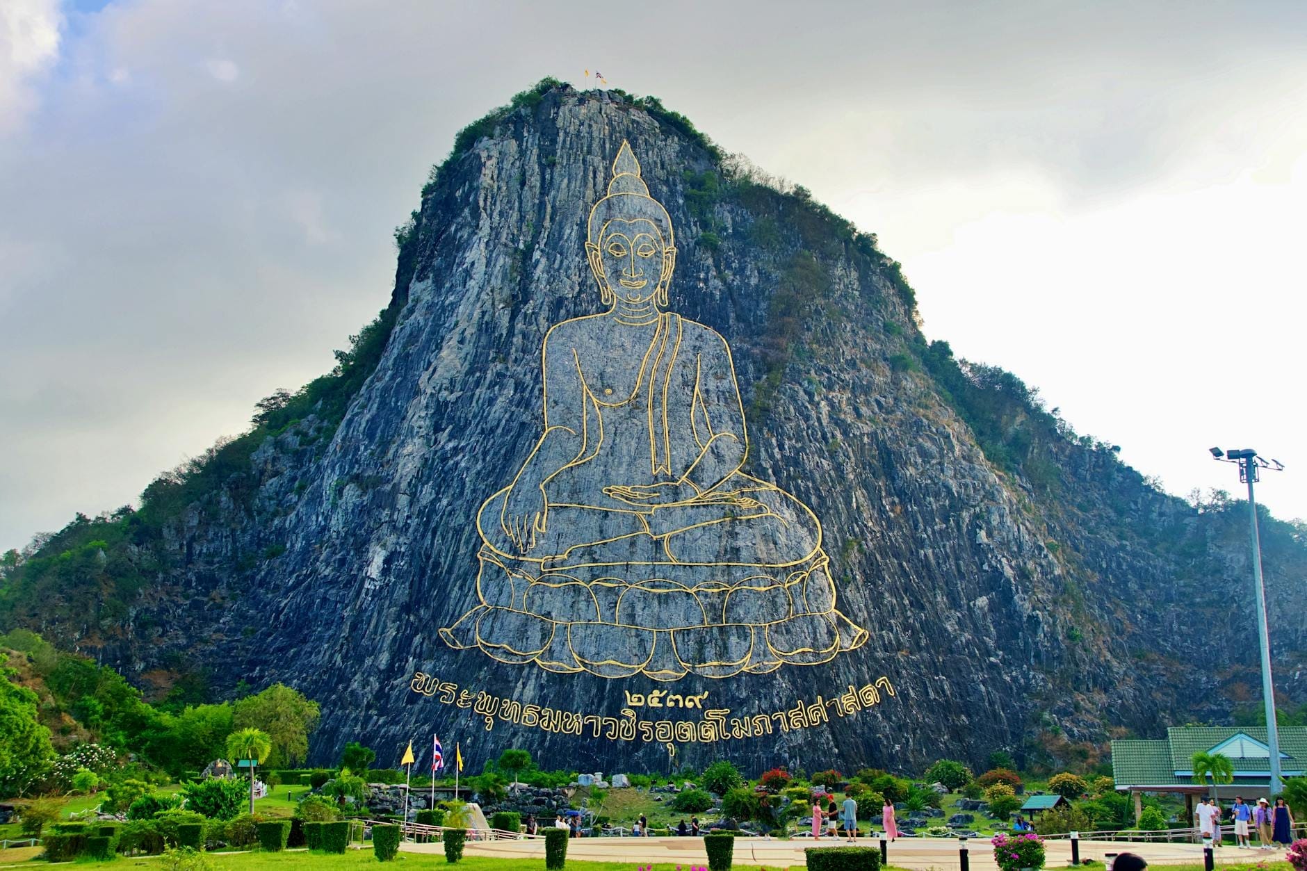 L'enorme immagine del Buddha delineata in oro e scolpita con il laser sulla montagna calcarea di Khao Chi Chan, circondata da giardini ordinati e alcuni visitatori in primo piano.