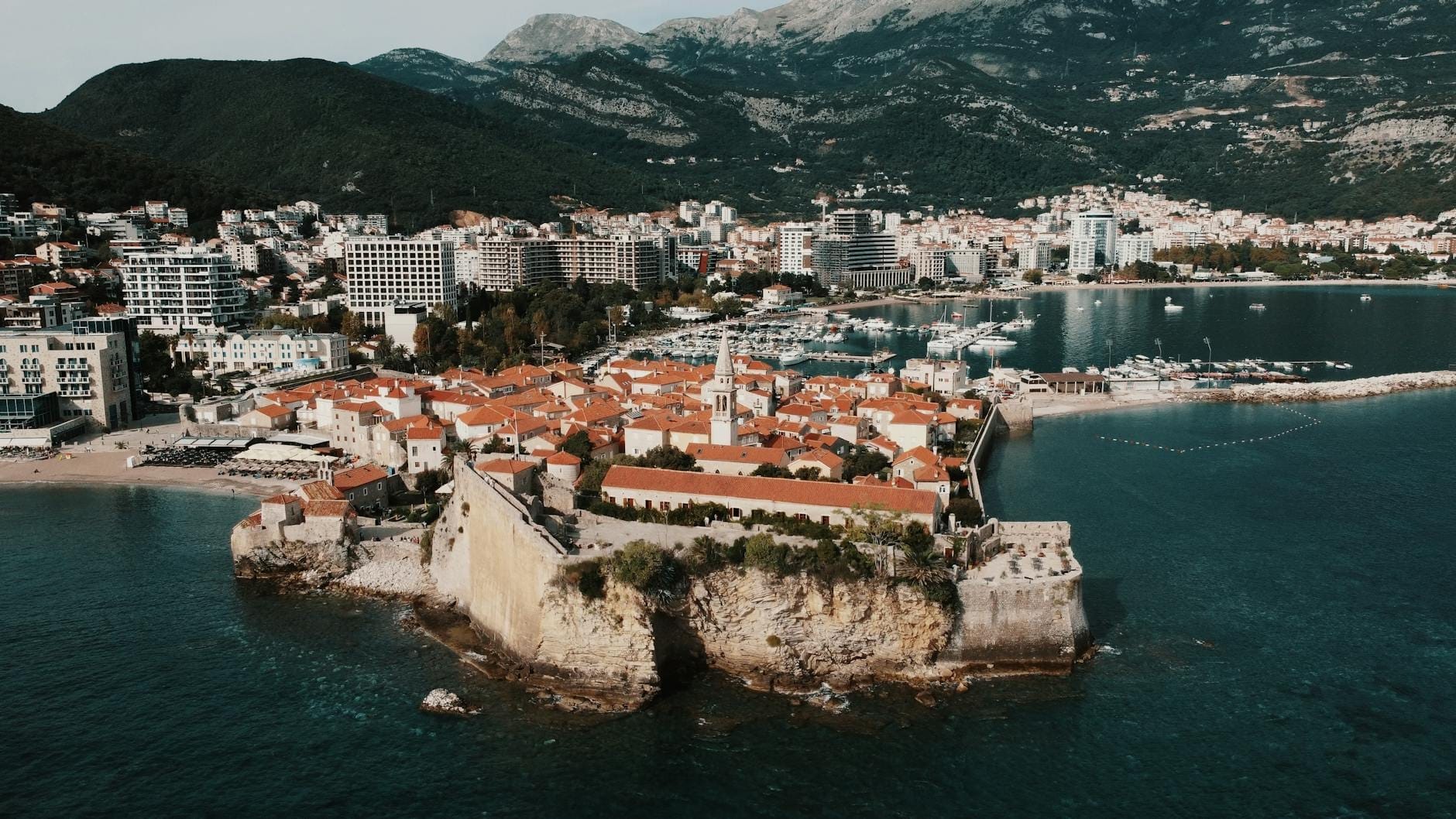 Vista aerea della Città Vecchia e della Cittadella di Budva sulla costa adriatica del Montenegro, con le mura medievali in pietra, i tetti rossi, il mare blu e le montagne sullo sfondo.