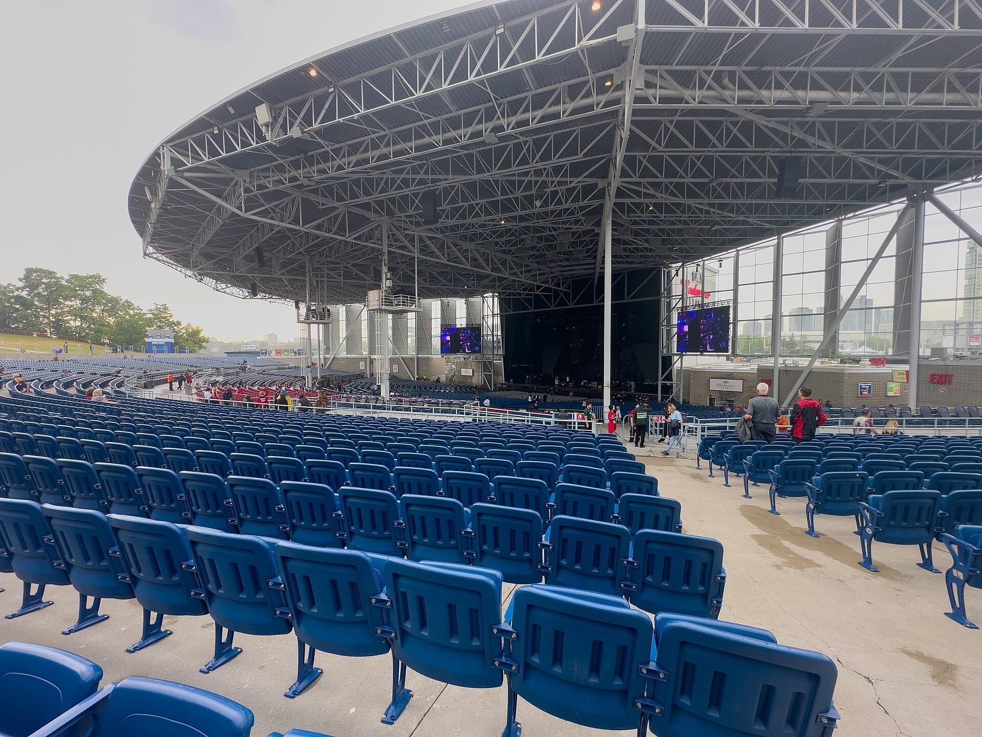 มุมมองกว้างของ RBC Amphitheatre ที่มีที่นั่งสีฟ้าเป็นชั้นๆ ภายใต้หลังคาเหล็กสมัยใหม่ พร้อมผู้คนบางส่วนที่มาชุมนุมก่อนคอนเสิร์ตริมน้ำ
