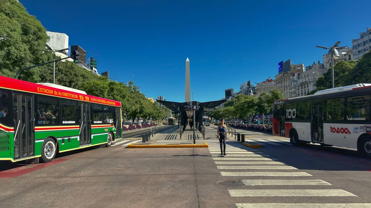 รถบัสและคนกำลังข้ามถนนที่ทางม้าลายบน Avenida 9 de Julio โดยมี Obelisco de Buenos Aires เป็นฉากหลัง