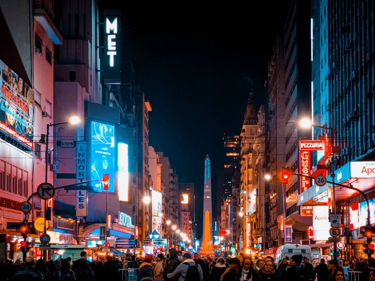Avenida Corrientes ที่แออัดในบัวโนสไอเรสยามค่ำคืน พร้อมป้ายนีออน ทางเท้าที่คึกคัก และ Obelisco ที่สว่างไสวอยู่เบื้องหลัง