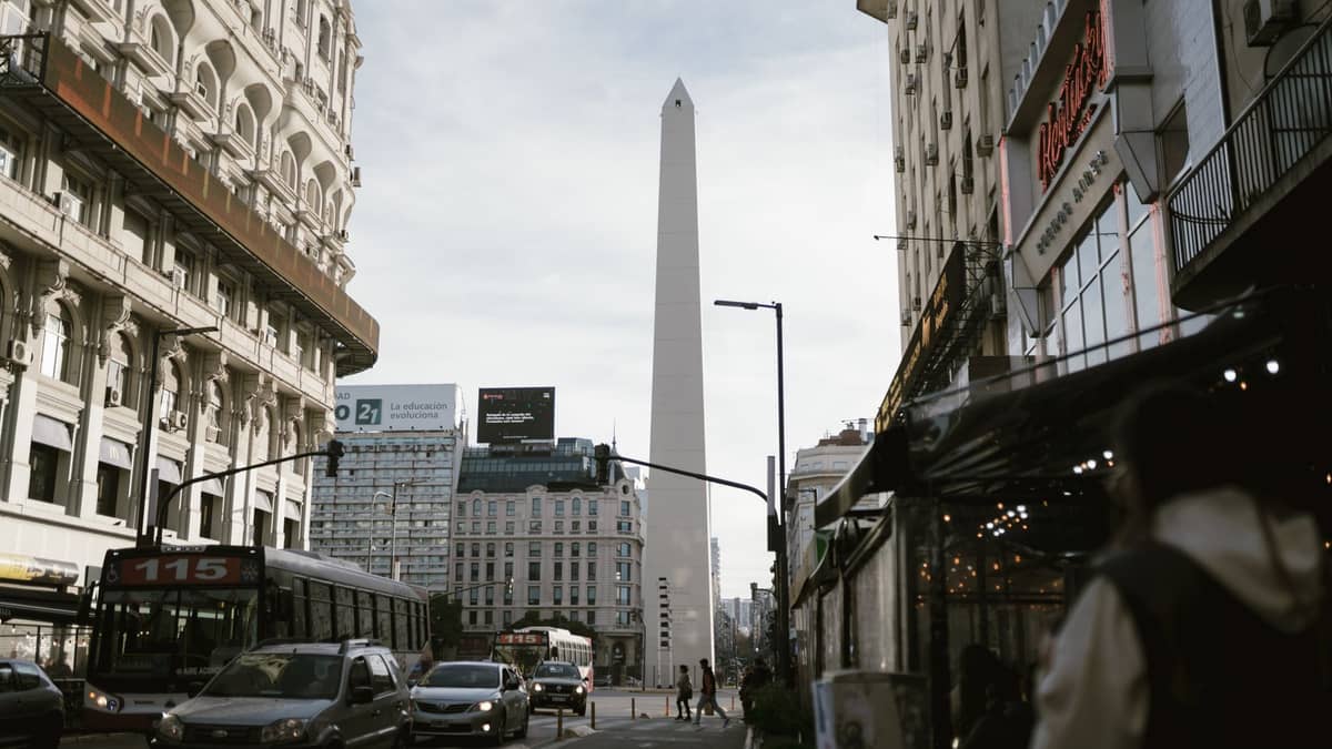ยอดObelisk แห่งบัวโนสไอเรสตระหง่านเหนือ Avenida Corrientes พร้อมฟุตปาธที่คึกคัก ร้านค้า และการจราจรที่สะท้อนบรรยากาศย่านช้อปปิ้งของเมือง