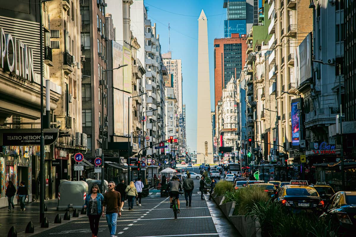 วิวระดับถนนที่คึกคักมุ่งสู่ Obelisco de Buenos Aires ซึ่งประกอบด้วยตึกสูงและผู้คนที่เดินอยู่บนทางเท้า