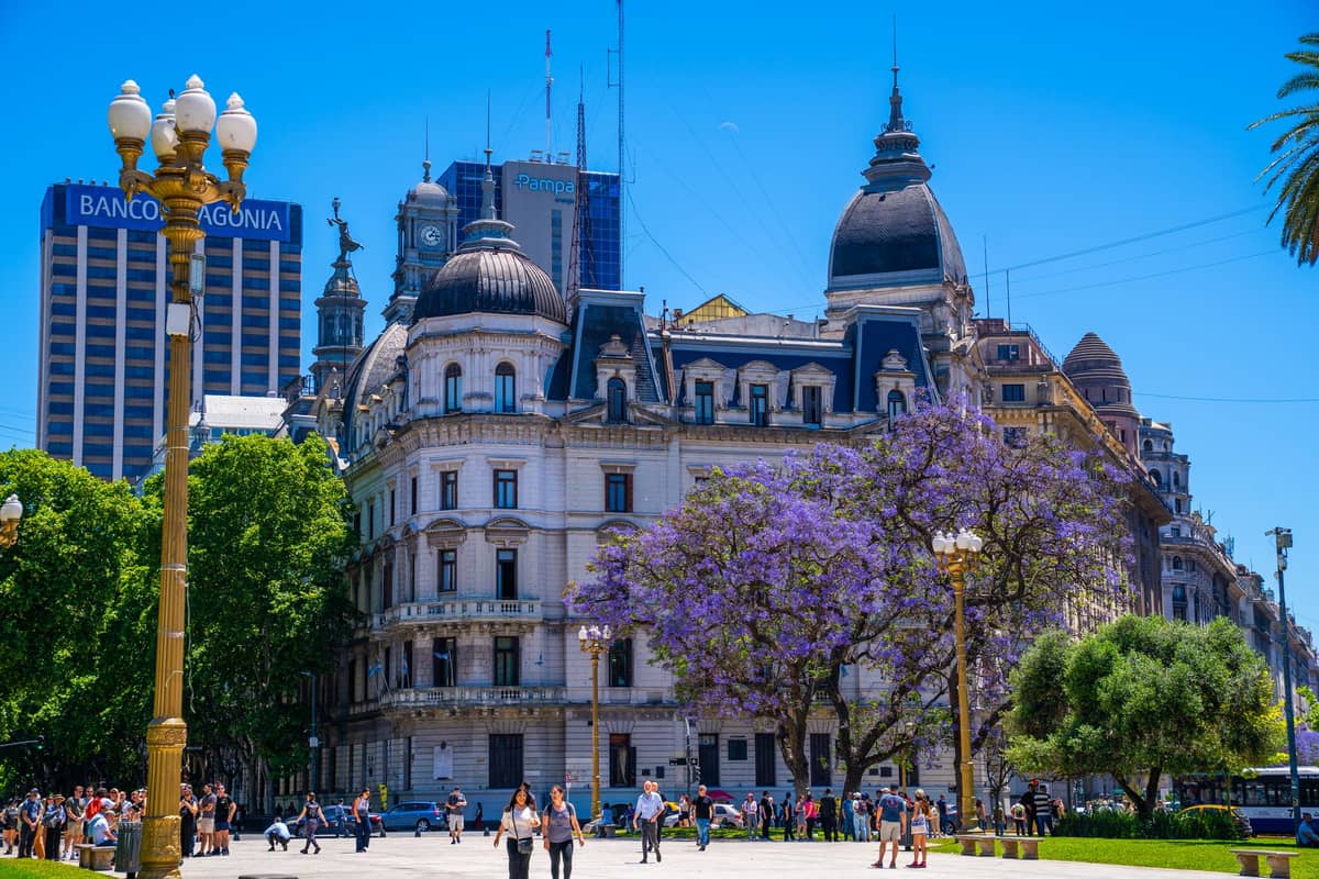 วิวกว้างของ Plaza de Mayo พร้อมสถาปัตยกรรมประดับประดาต้นศตวรรษที่ 20 ต้นจาการันดา ผู้คนเดินเล่น และอาคารเมืองในย่านประวัติศาสตร์ของบัวโนสไอเรส