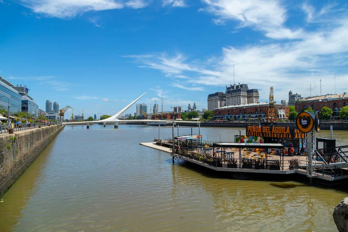วิวกว้างของพื้นที่ริมน้ำ Puerto Madero ในบัวโนสไอเรส พร้อมอาคารสมัยใหม่ สะพานอันเป็นสัญลักษณ์ และผู้คนที่เดินเล่นริมแม่น้ำใต้ท้องฟ้าสีฟ้าแจ่มใส
