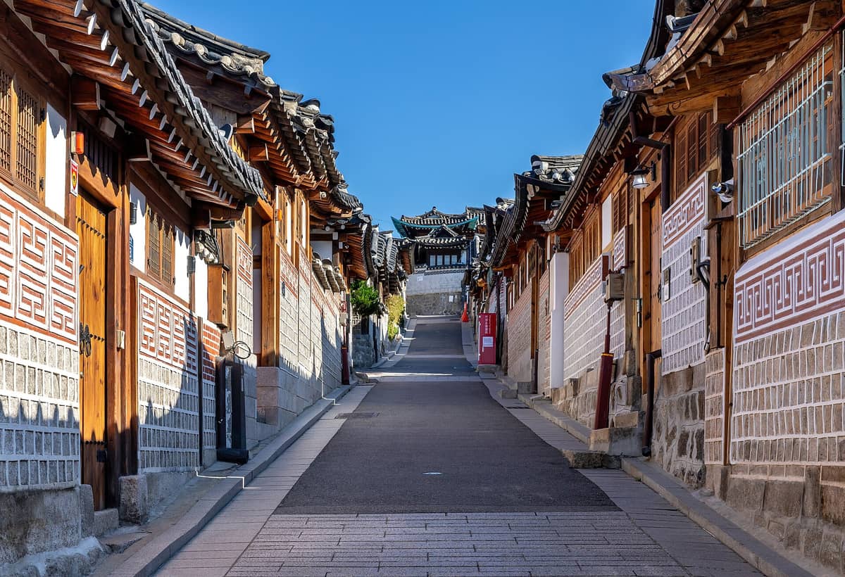 Vista ampla de uma viela tradicional de pedra e tijolo ladeada por casas hanok em Bukchon Hanok Village sob um céu azul bem aberto.