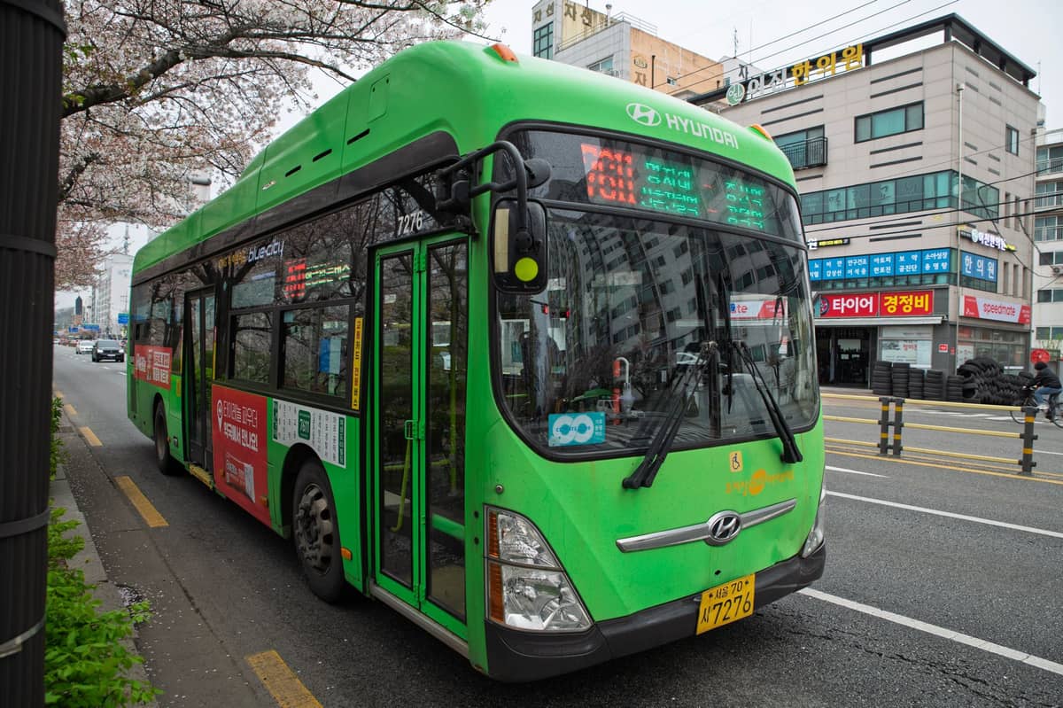 Um ônibus urbano verde estacionado em uma rua de Seul com placas em coreano e edifícios ao fundo, típico do transporte público local.
