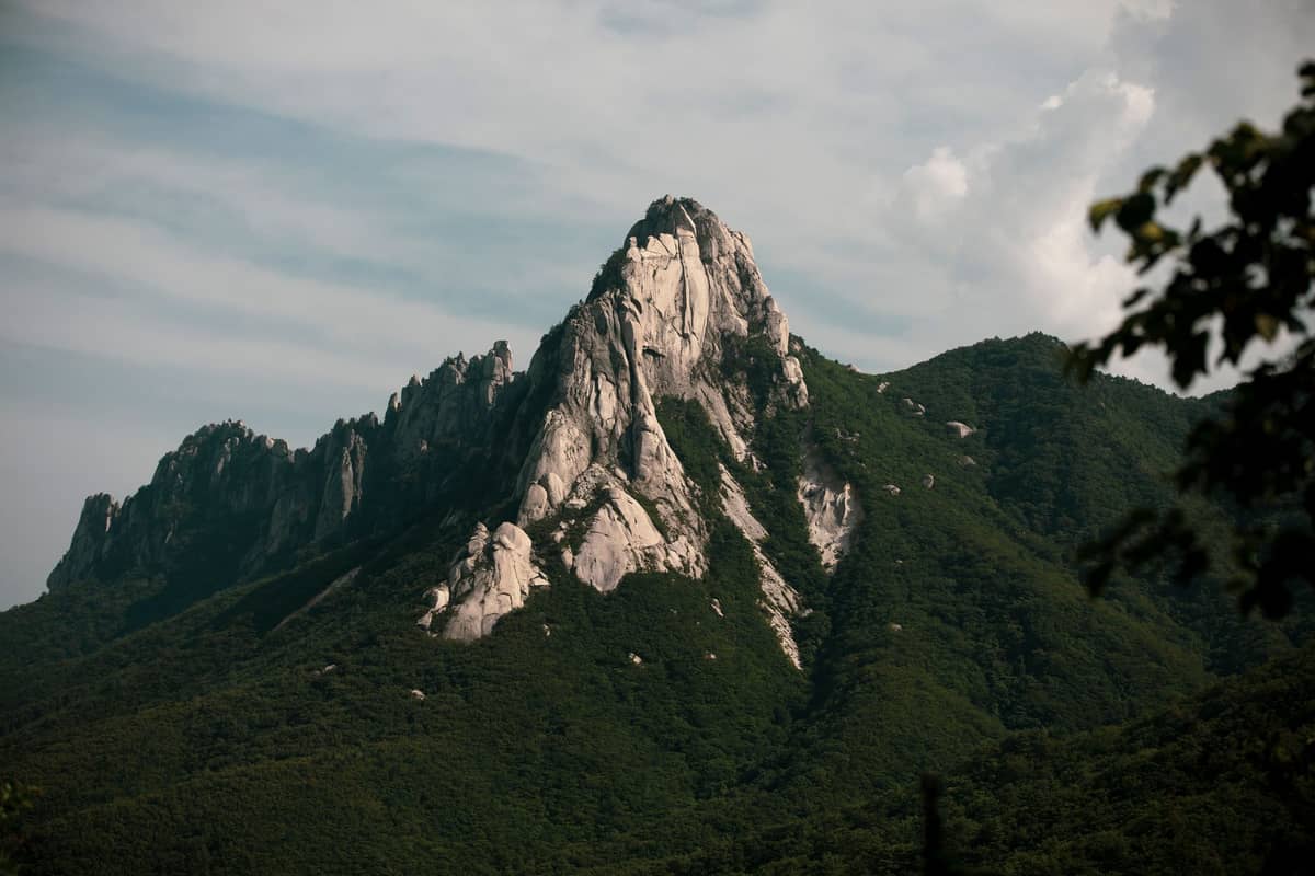 Picos irregulares de granito de Bukhansan erguendo-se acima de encostas arborizadas verdes sob um céu parcialmente nublado, sem pessoas visíveis.