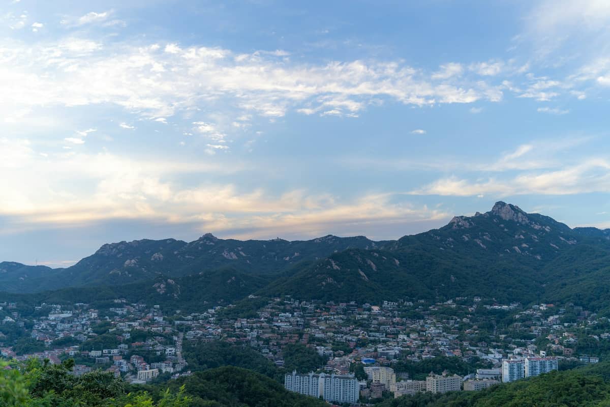 Vista panorâmica dos picos da montanha Bukhansan emergindo acima dos bairros do norte de Seul sob um céu azul com nuvens esparsas.