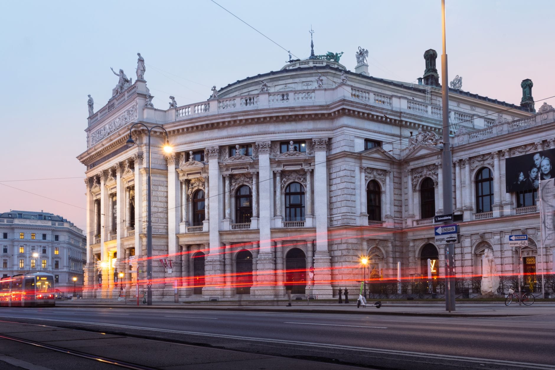 Das Burgtheater in der Abenddämmerung: Die Neorenaissance-Fassade an der Wiener Ringstraße ist beleuchtet, Straßenlaternen und vorbeifahrende Autos zeichnen dynamische Lichtspuren vor dem Gebäude.