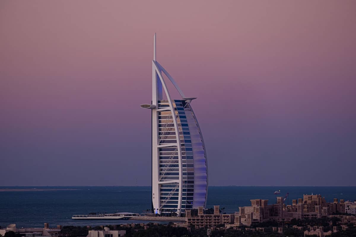 Das Burj Al Arab Hotel erhebt sich in der Abenddämmerung über der Küstenlinie – seine ikonische Segelform hebt sich deutlich vor einem violett-rosafarbenen Himmel und dem Meer im Hintergrund ab.