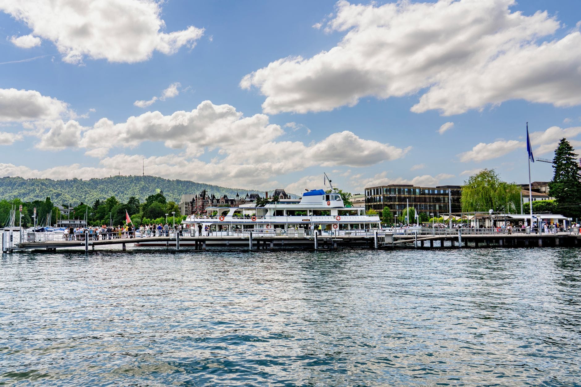 ภาพวิวมุมกว้าง Bürkliplatz และท่าเรือในซูริก เห็นเรือผู้โดยสารเทียบท่า ผู้คนเดินริมทาง น้ำ อาคารเมือง และเนินเขาอยู่ฉากหลัง