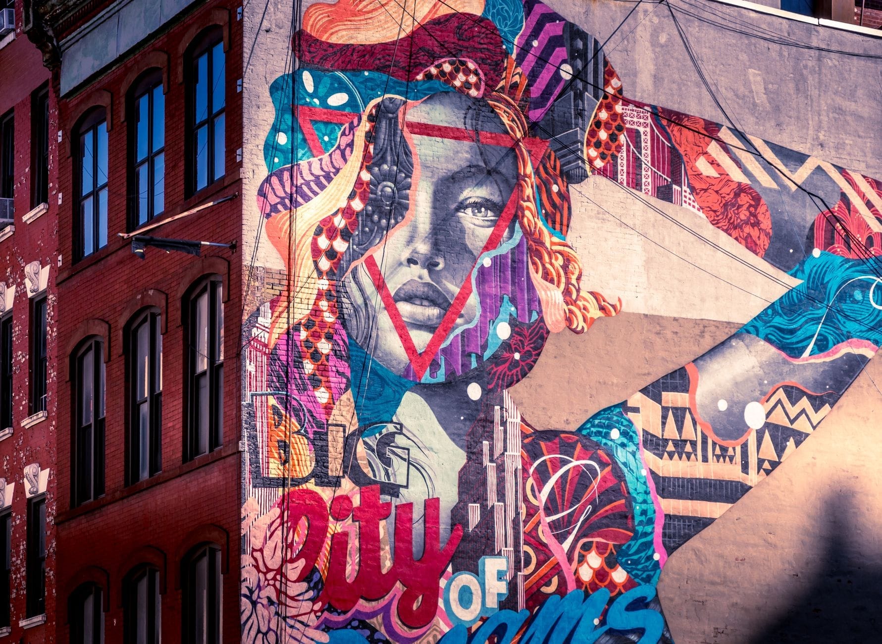 Grande murale colorato con il volto di una donna e la scritta 'Big City of Dreams' dipinta su un edificio in mattoni in un contesto urbano.