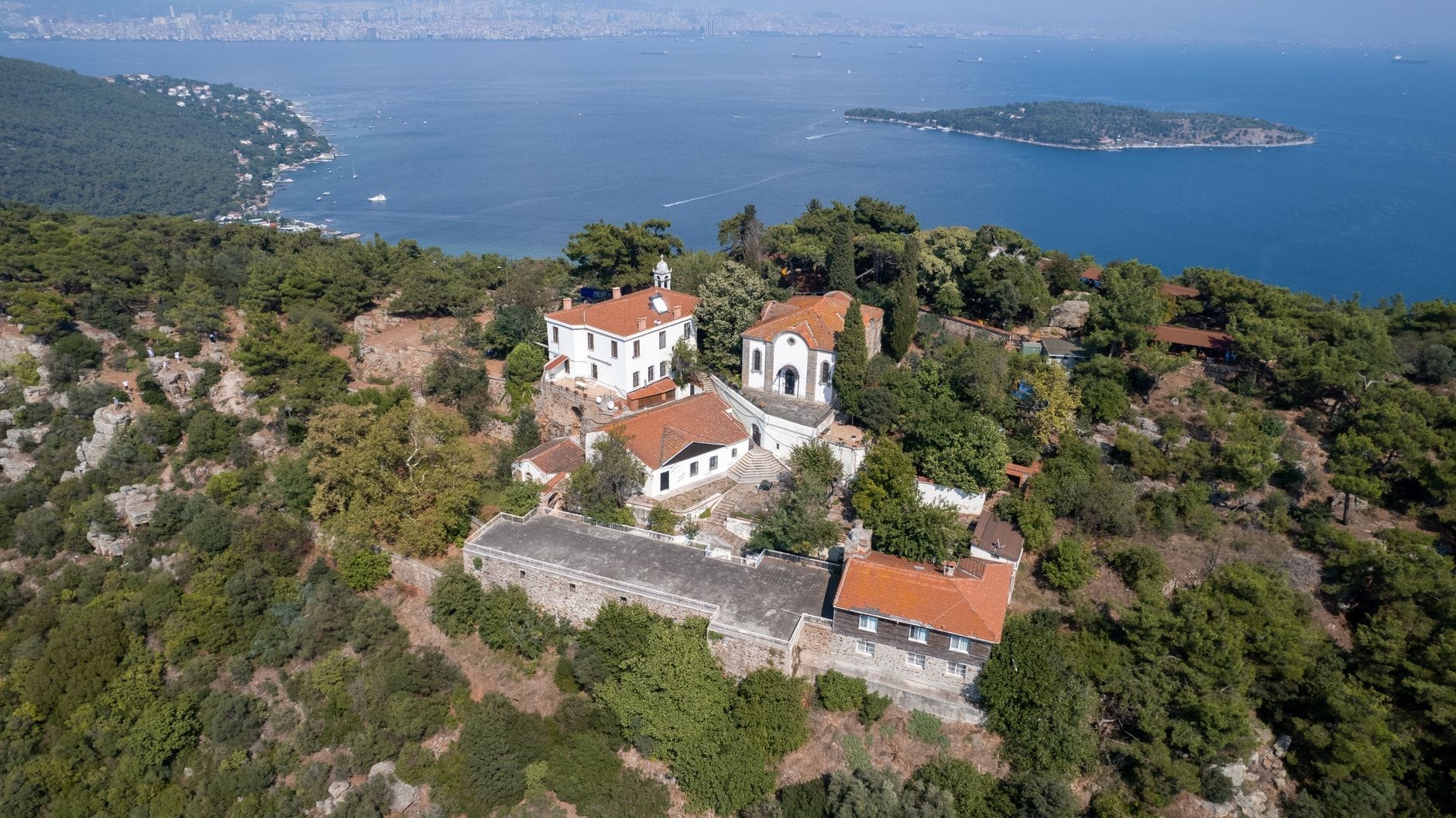 Vista aerea di Büyükada con storiche ville in legno, una piccola chiesa, rigogliosi pini e il blu profondo del Mar di Marmara, con Istanbul appena visibile sullo sfondo.