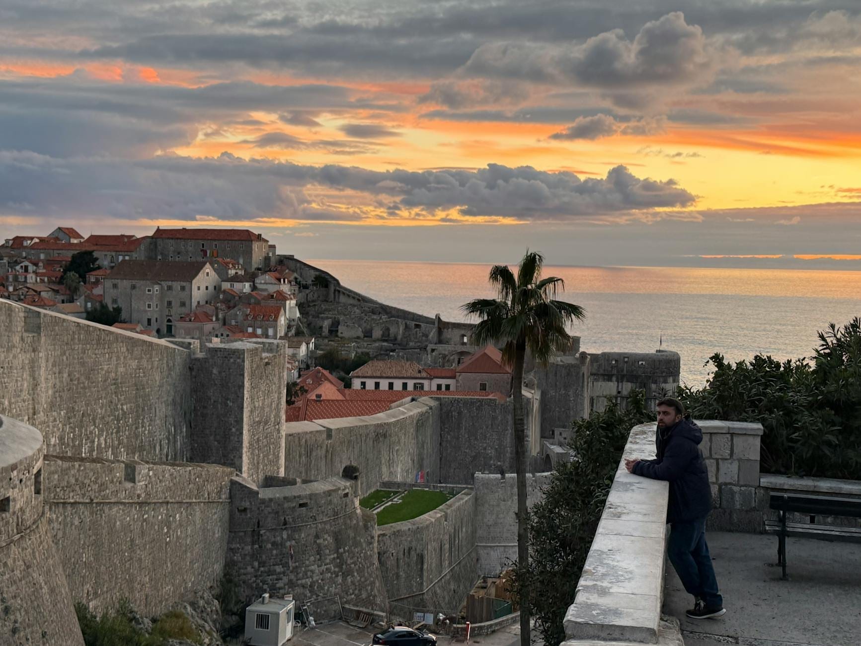 Vue des anciennes murailles de Dubrovnik au coucher du soleil, avec le soleil partiellement masqué par le mur et un homme regardant la mer.